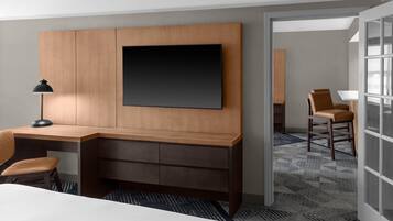 Habitación de hotel con un escritorio de madera, un televisor de pantalla plana montado en la pared, una silla, una lámpara y una alfombra estampada.