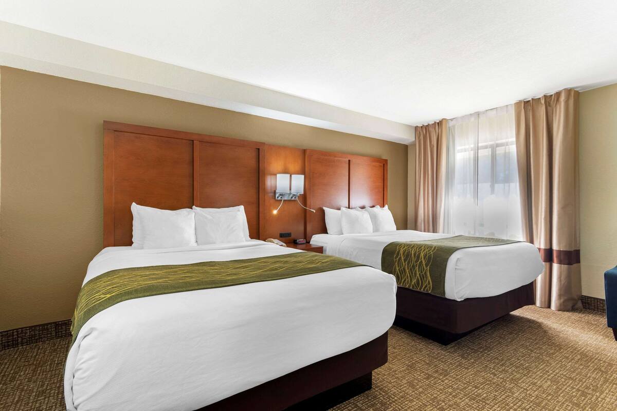 A hotel room with two beds, a wooden headboard, a nightstand, and a window with curtains.