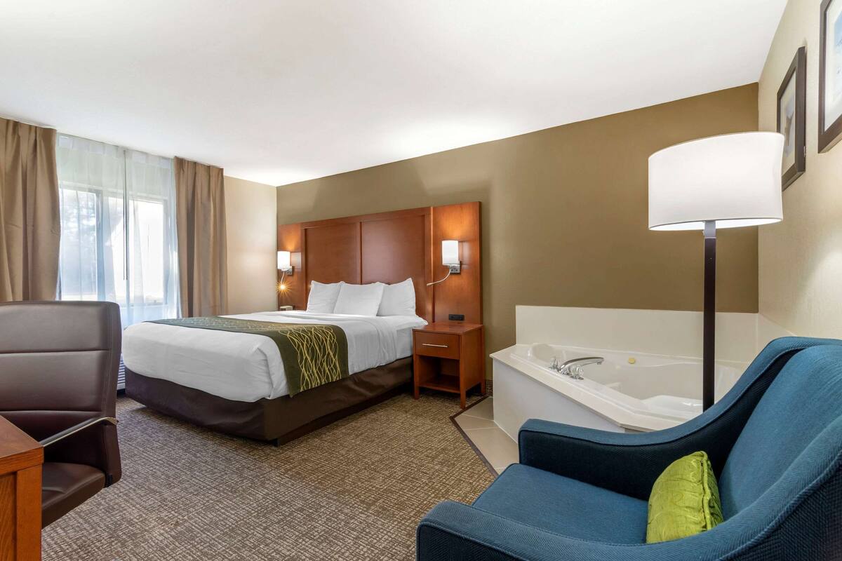 A hotel room with a bed, a desk, a chair, a bathtub, and a window with curtains.