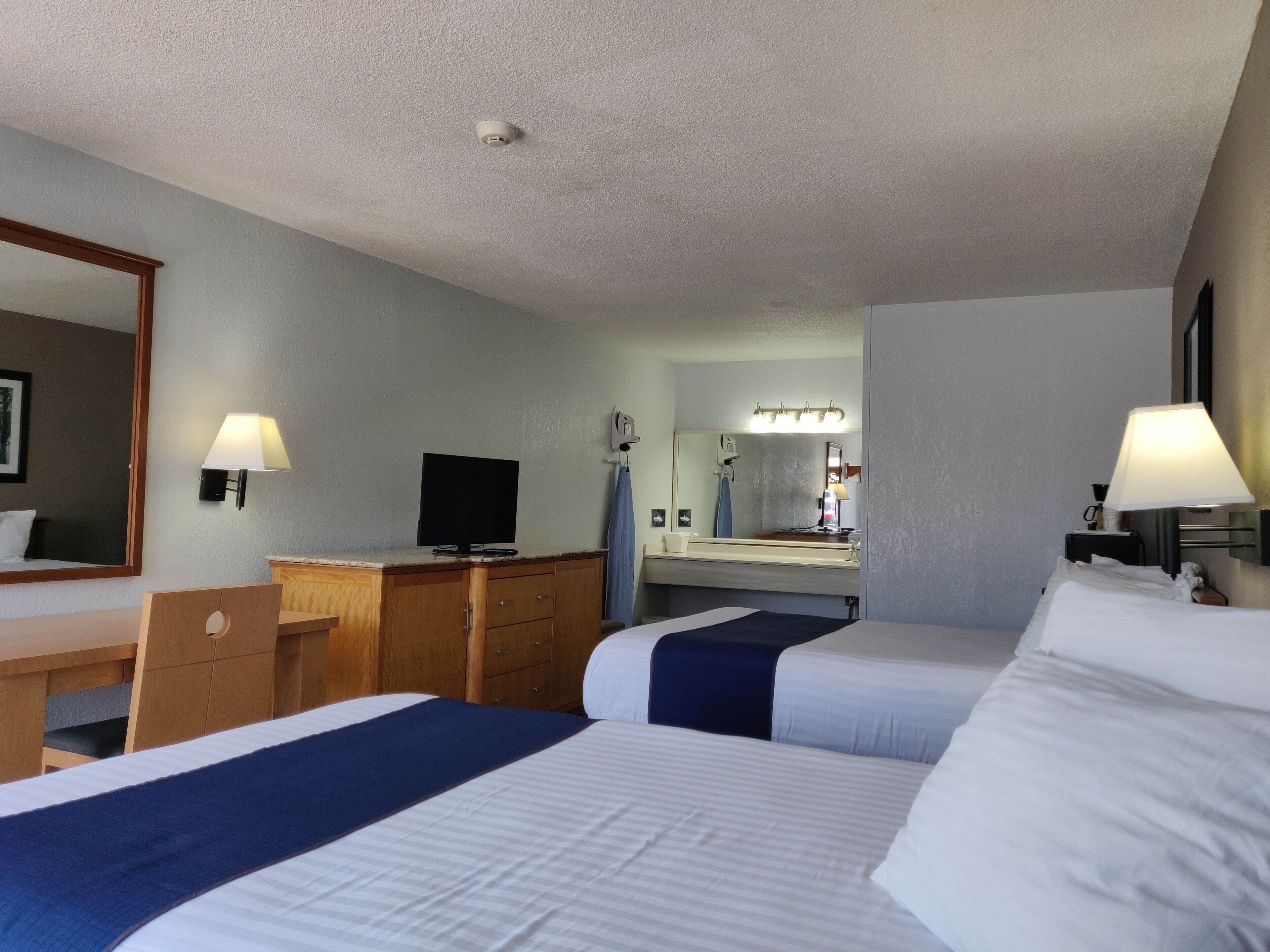 A hotel room with two beds, a desk, a television, a mirror, and a bathroom visible in the background.