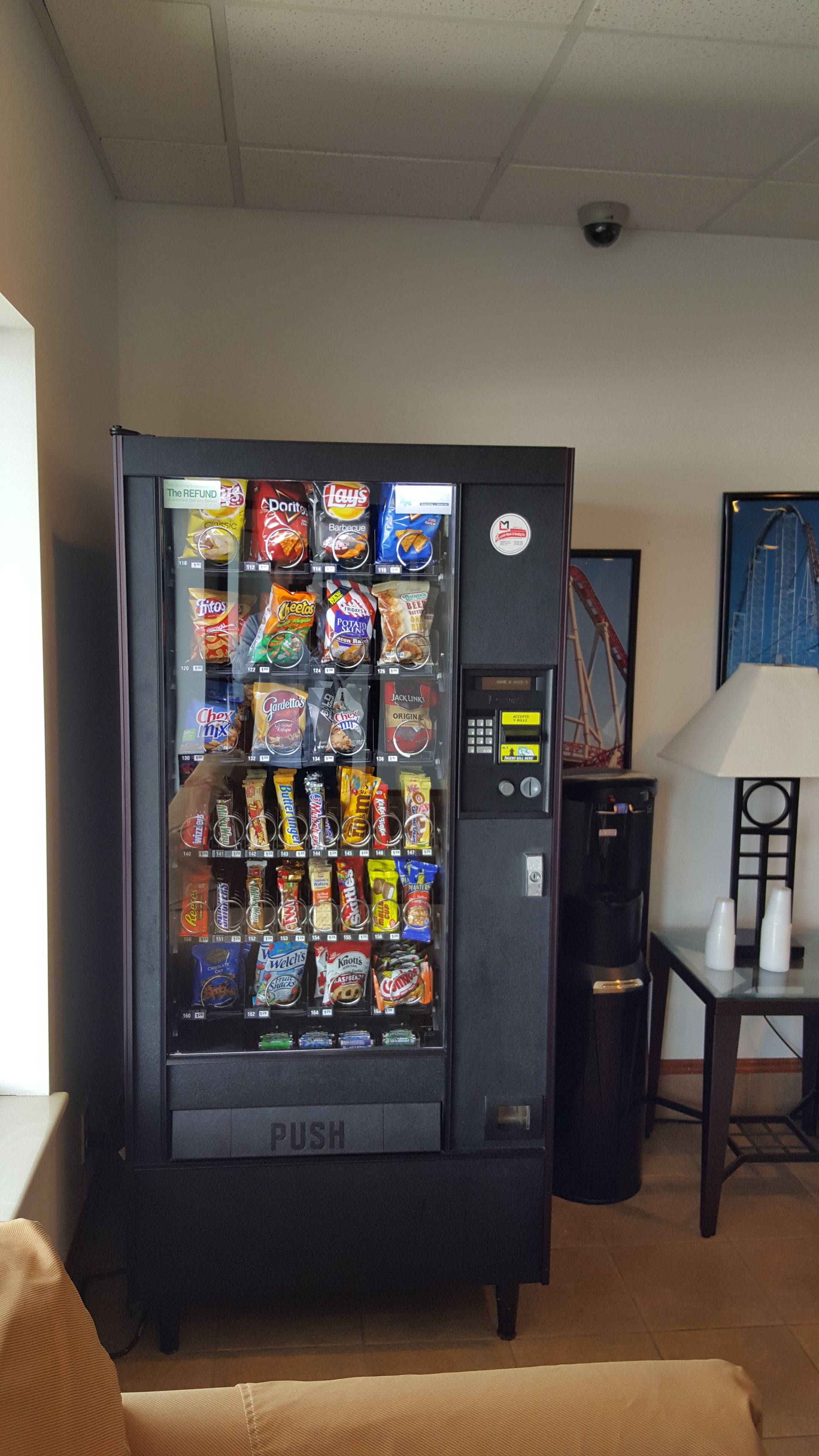 Máquina expendedora con aperitivos y bebidas, ubicada en una habitación con una lámpara y un cuadro en la pared.