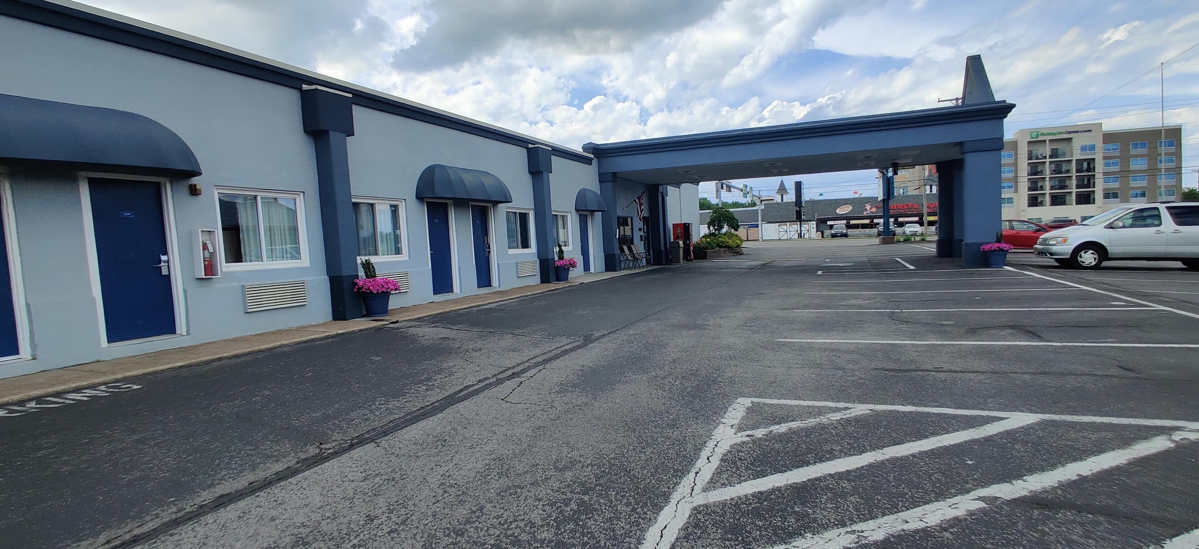 A motel with a row of rooms under a covered walkway, a parking lot, and a building in the background.