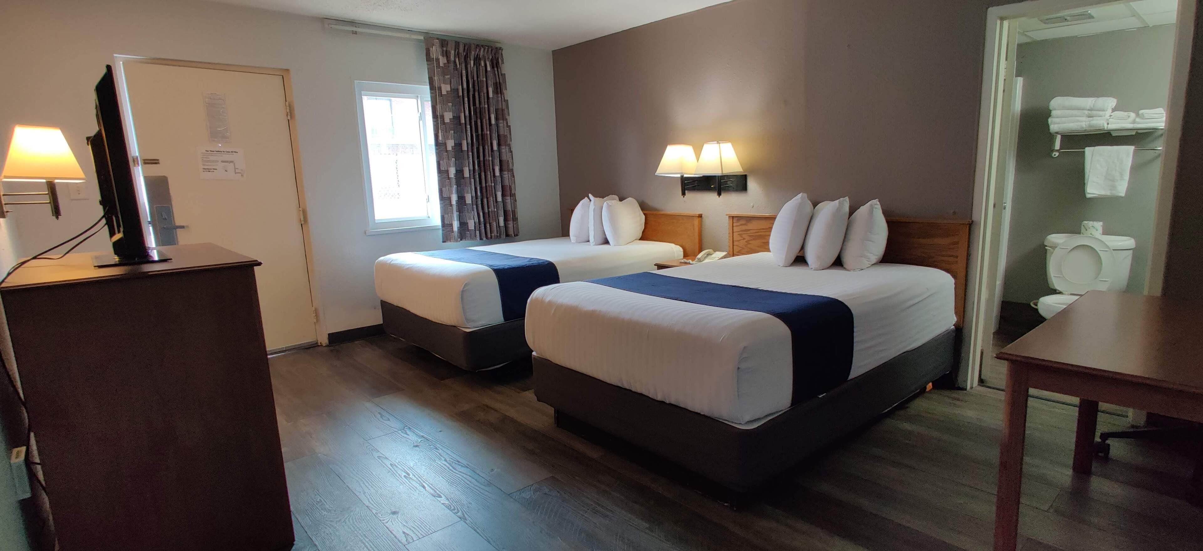 A hotel room with two single beds, a desk, a lamp, a window with curtains, and a bathroom visible through an open door.