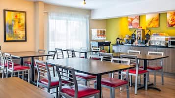 Un comedor moderno con varias mesas y sillas, una encimera de cocina con electrodomésticos y un ventanal con cortinas translúcidas.