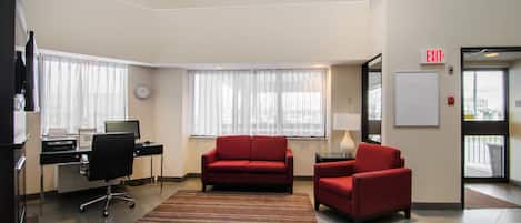 A modern hotel room with a desk, computer, red sofa, and a view of the city through large windows.