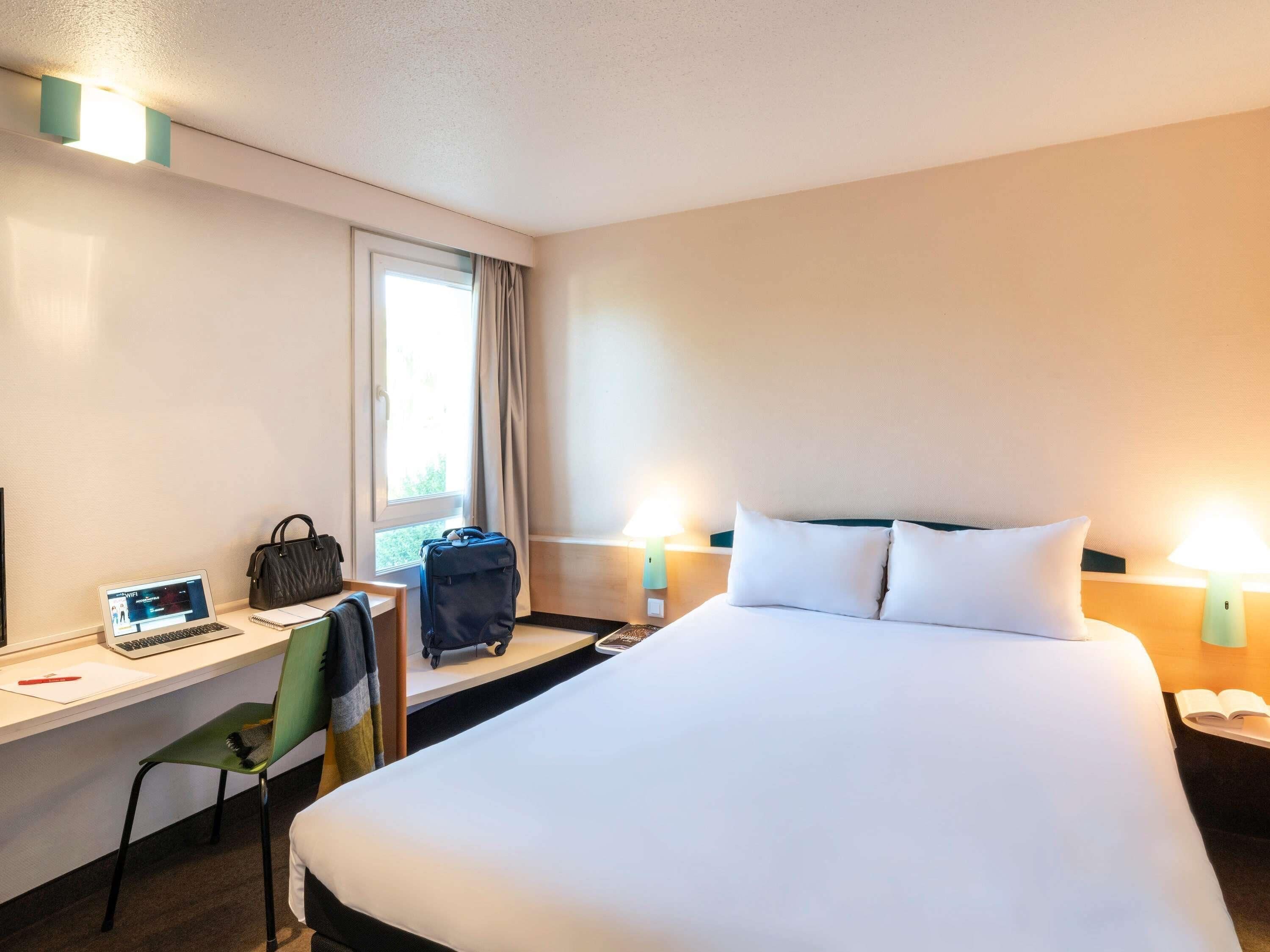 Ein Hotelzimmer mit Bett, Schreibtisch, Stuhl, Laptop, Handtasche und Koffer.