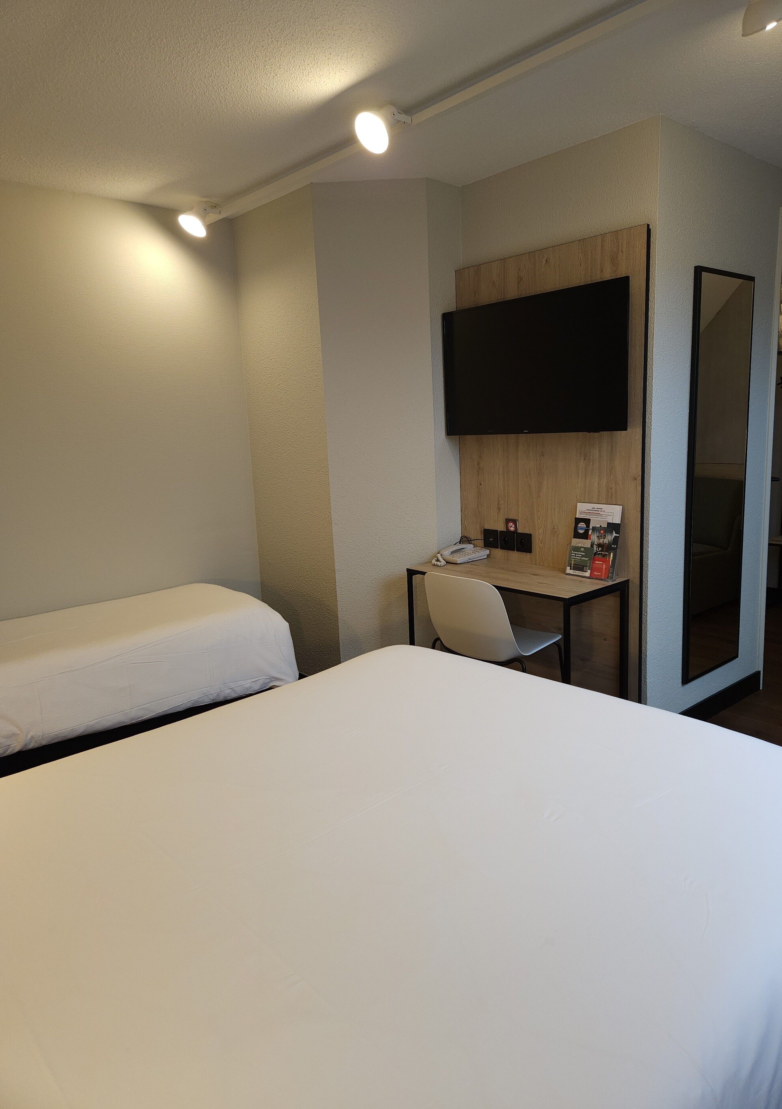 A hotel room with two single beds, a desk with a chair, a flat-screen TV mounted on the wall, and a mirror.