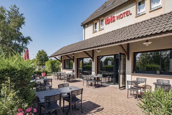 Un patio al aire libre con mesas y sillas, una zona de estar techada y un edificio con un letrero que dice “ibis HOTEL”.