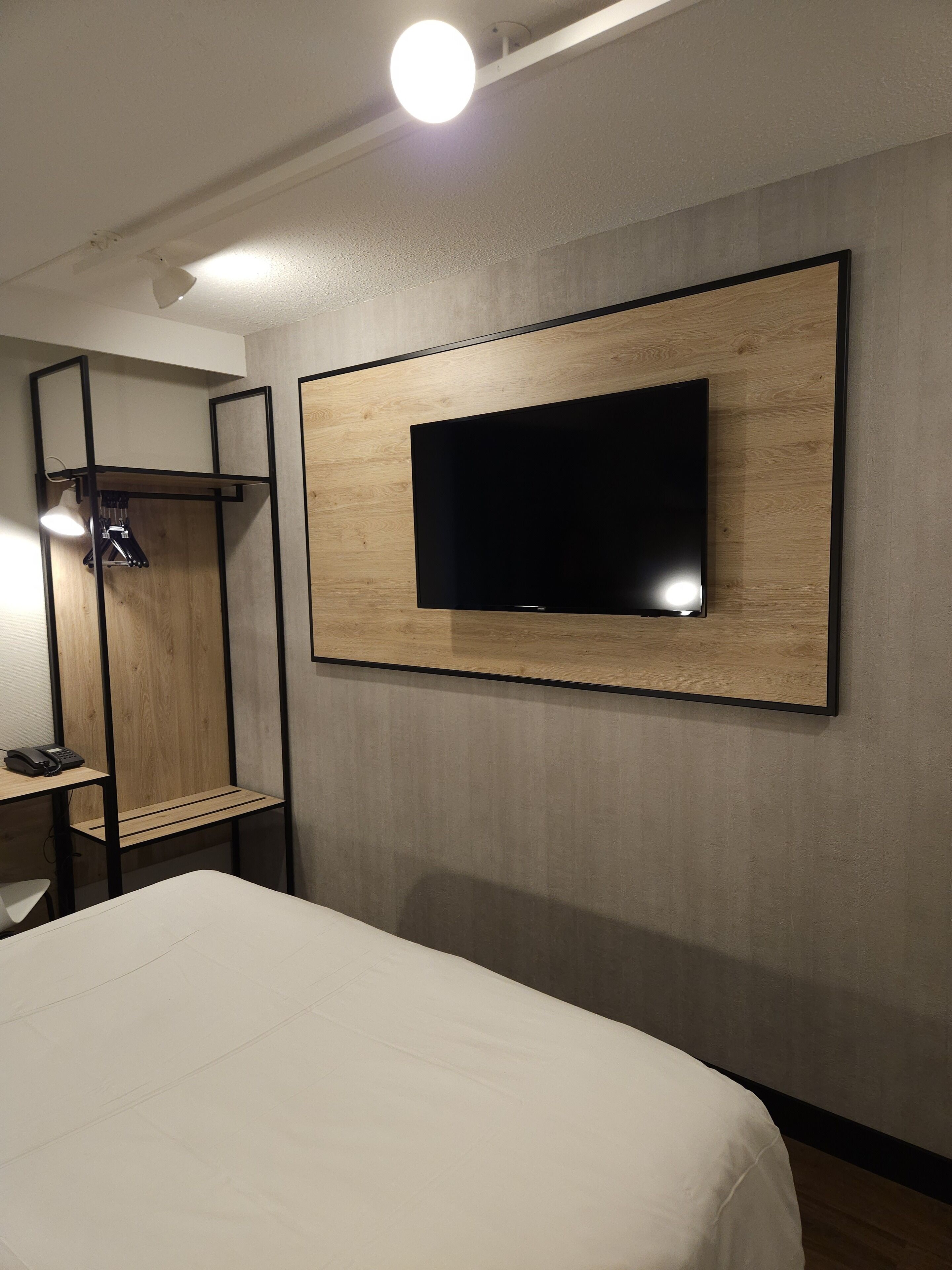 A hotel room with a bed, a mounted TV, a desk with a chair, and a wardrobe.
