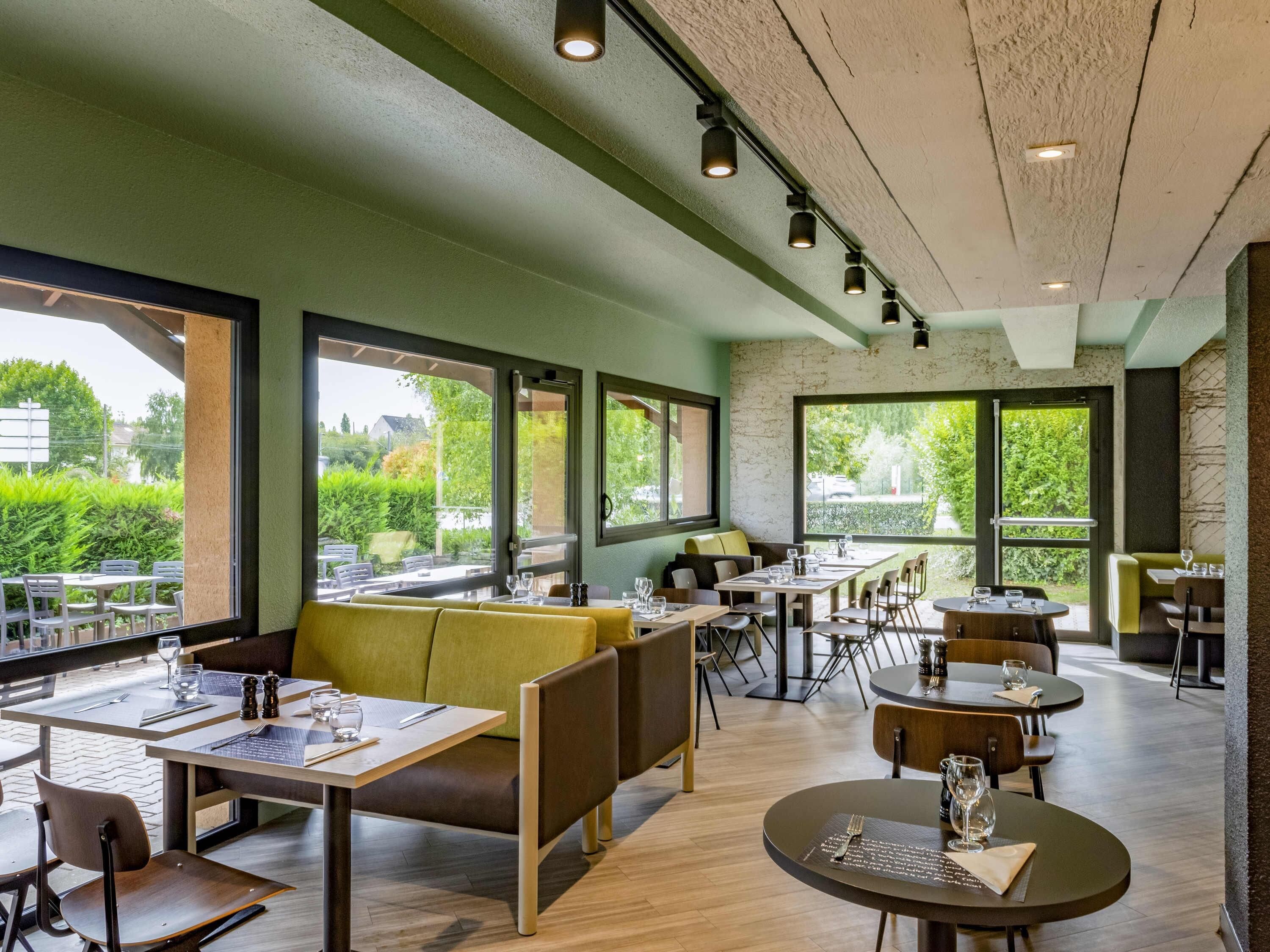 Un restaurante con mesas en el interior, amplias ventanas y vistas a zonas verdes.