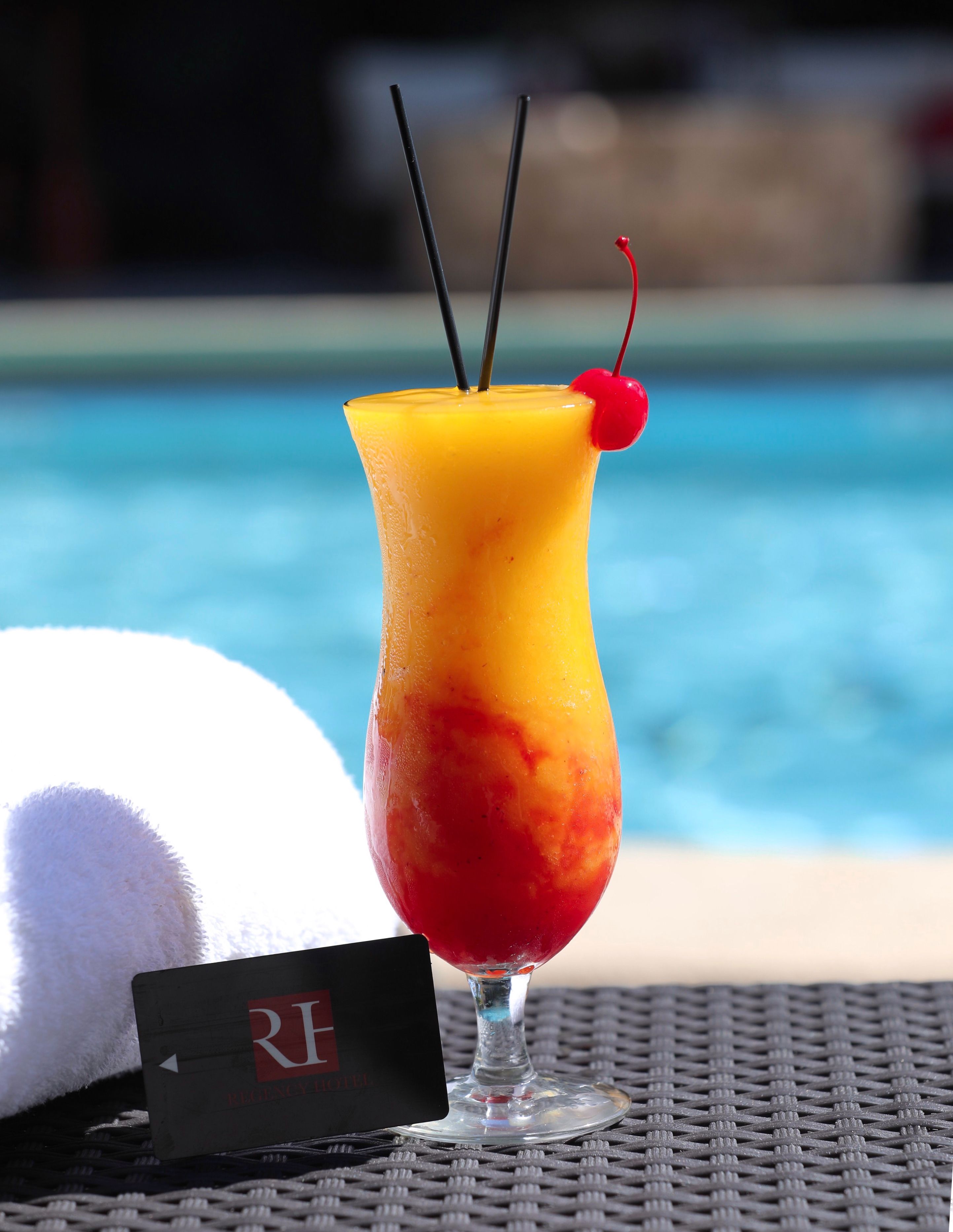 En drink med en färgskala, ett körsbär som dekoration och två sugrör, placerad på ett bord med en pool i bakgrunden.