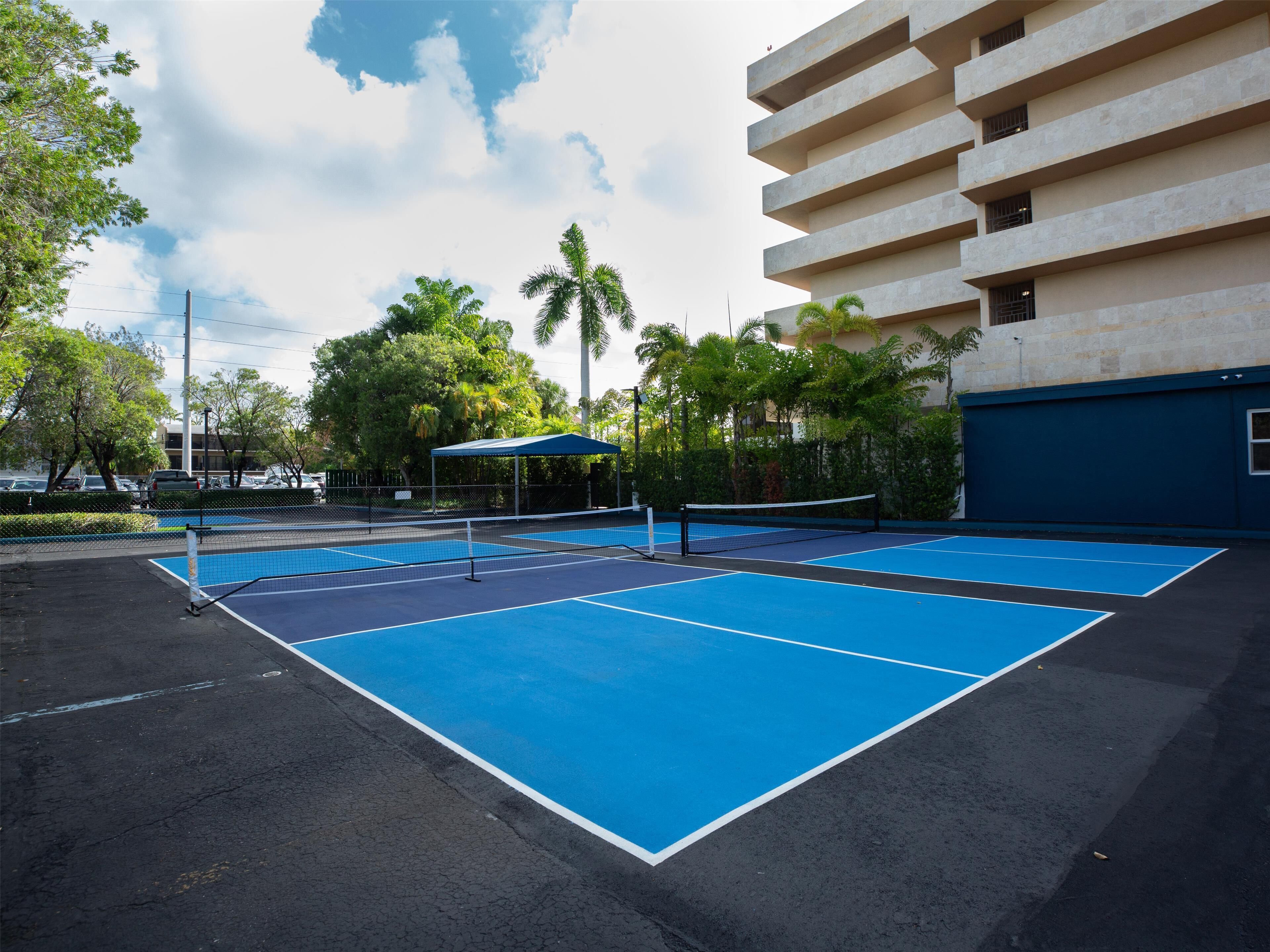Una cancha de tenis al aire libre con red, rodeada de árboles y un edificio.