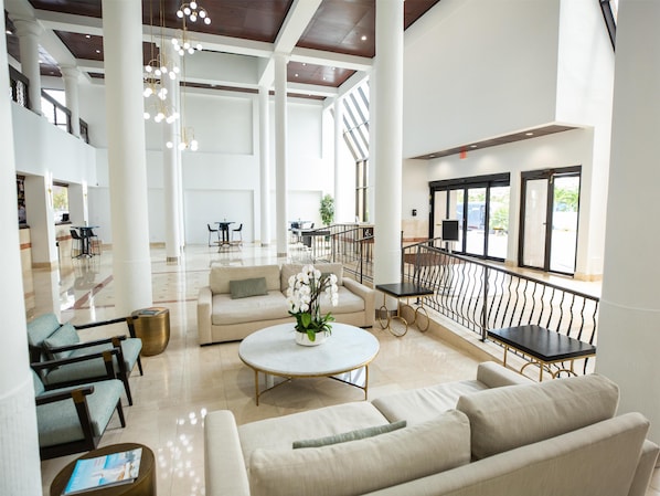 A spacious lobby with high ceilings, large windows, and a central seating area with a round table and sofas.