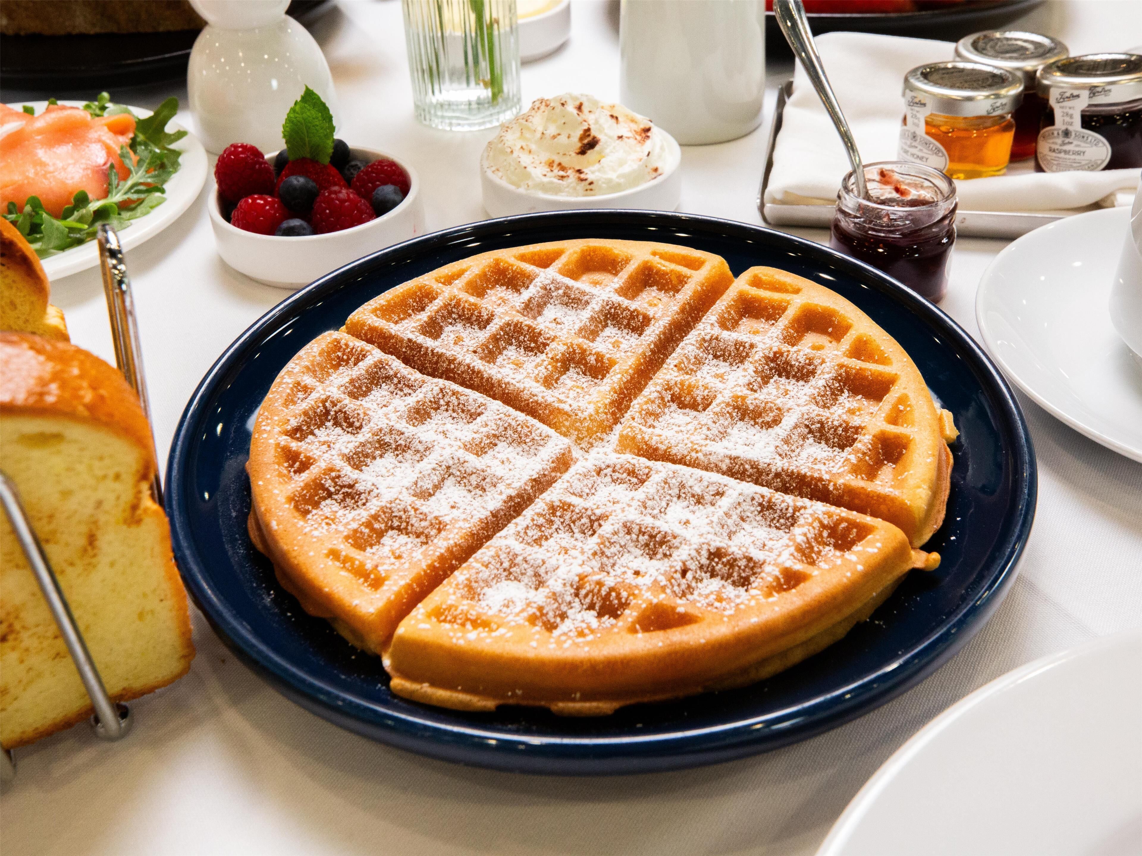 Um prato de waffles recém-feitos com açúcar de confeiteiro, acompanhado de uma tigela com frutas vermelhas e um pequeno recipiente de chantilly.