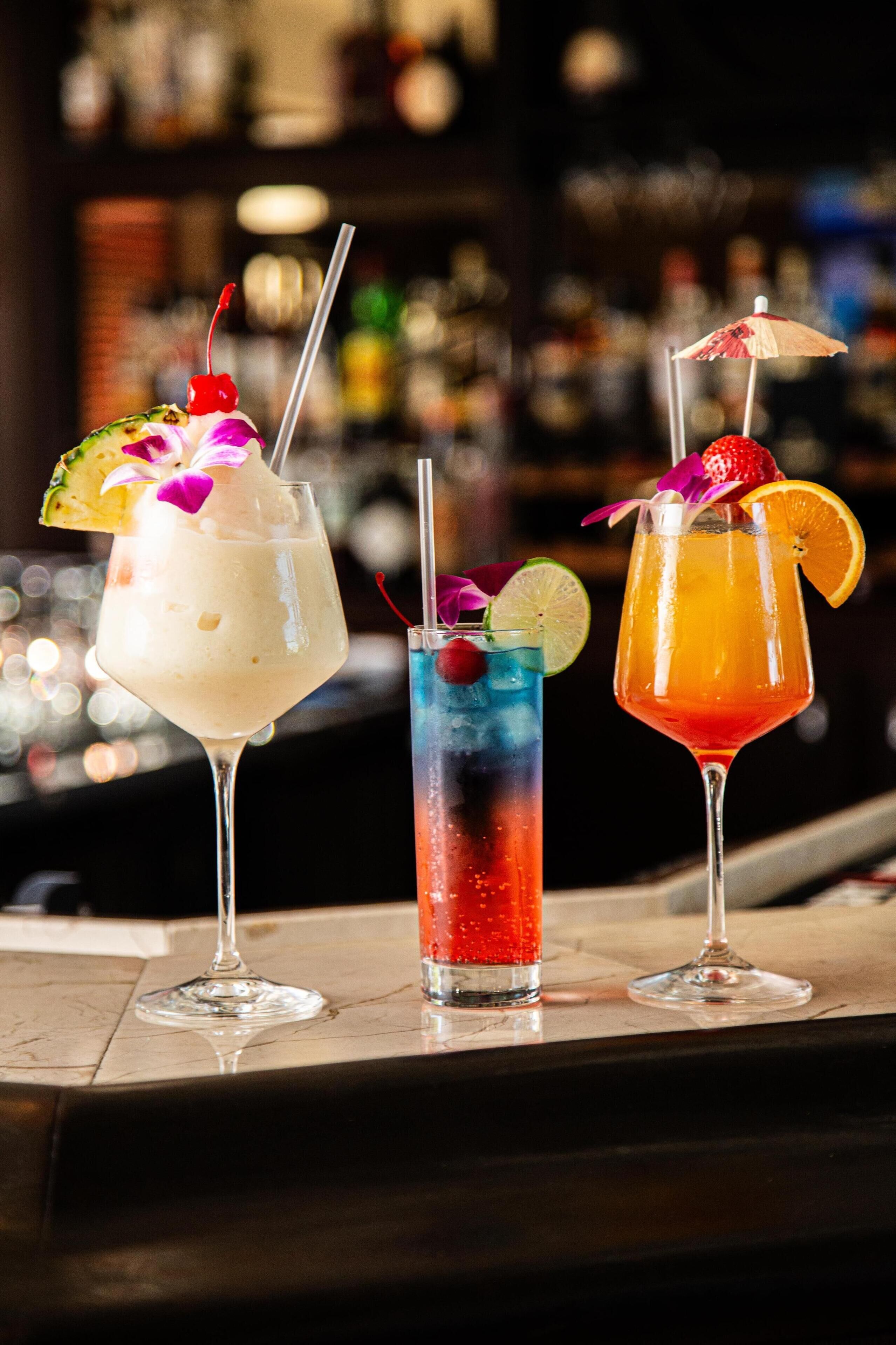 Three cocktails on a bar counter with a blurred background of bottles.