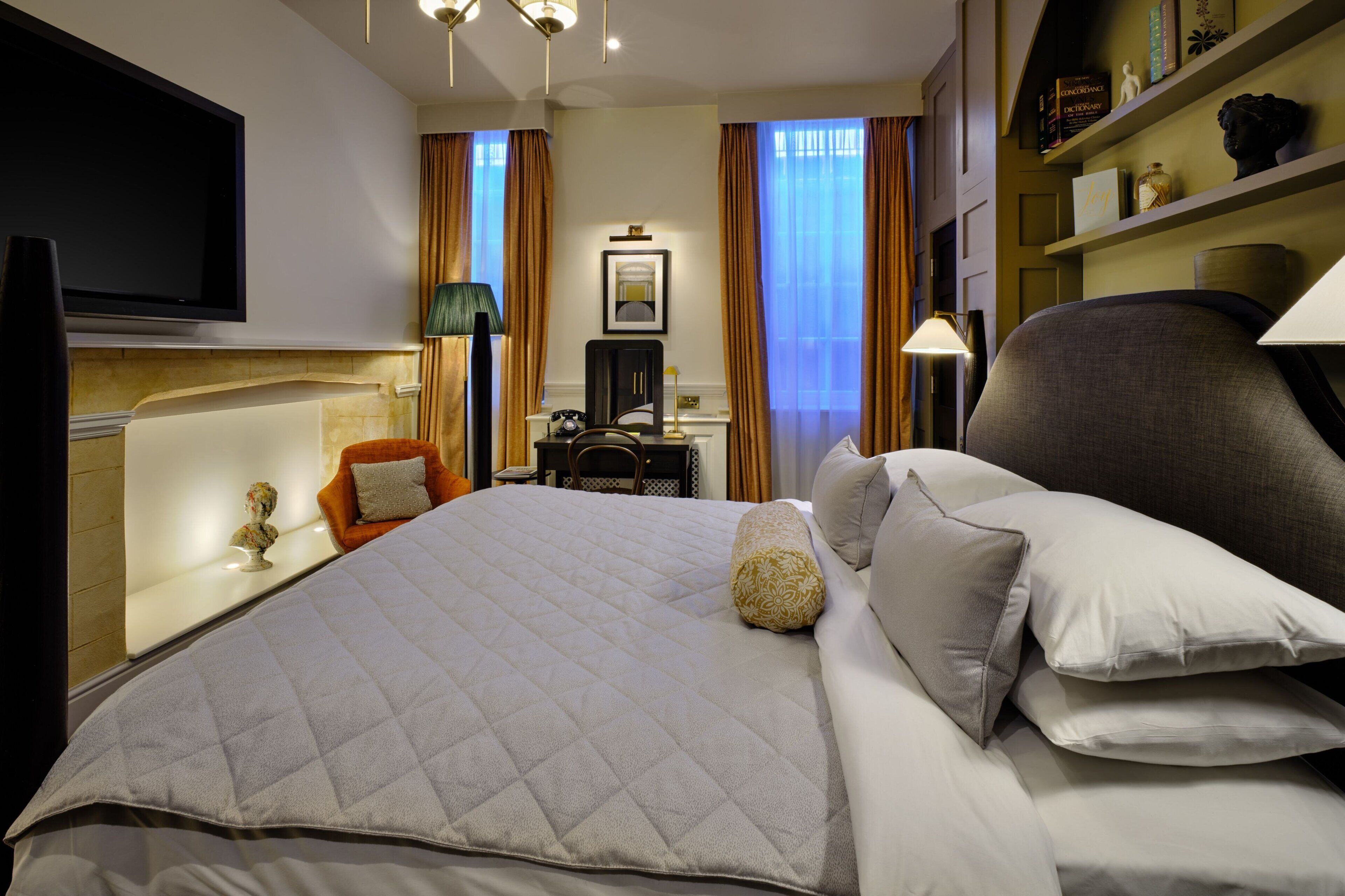 A hotel room with a large bed, a flat-screen TV mounted on the wall, a desk with a chair, and a bookshelf with decorative items.