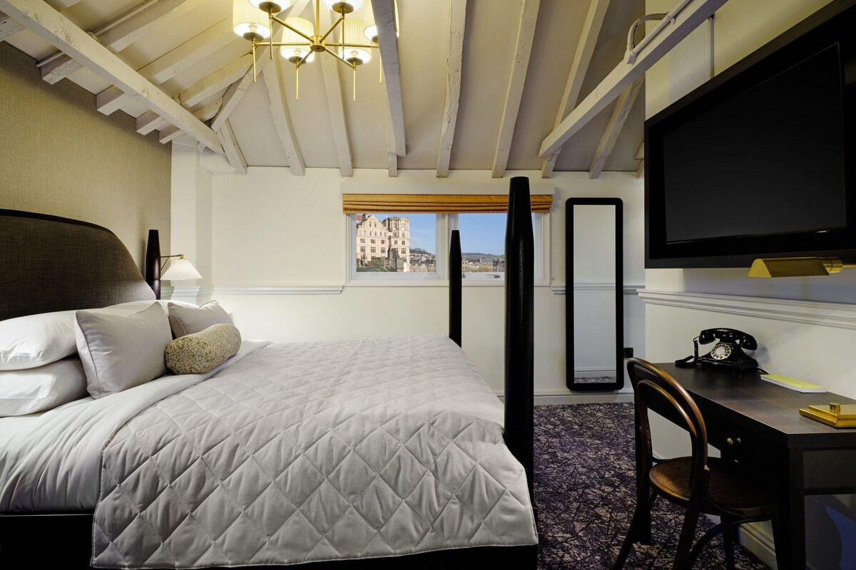A hotel room with a bed, a desk with a telephone, a large mirror, and a TV.