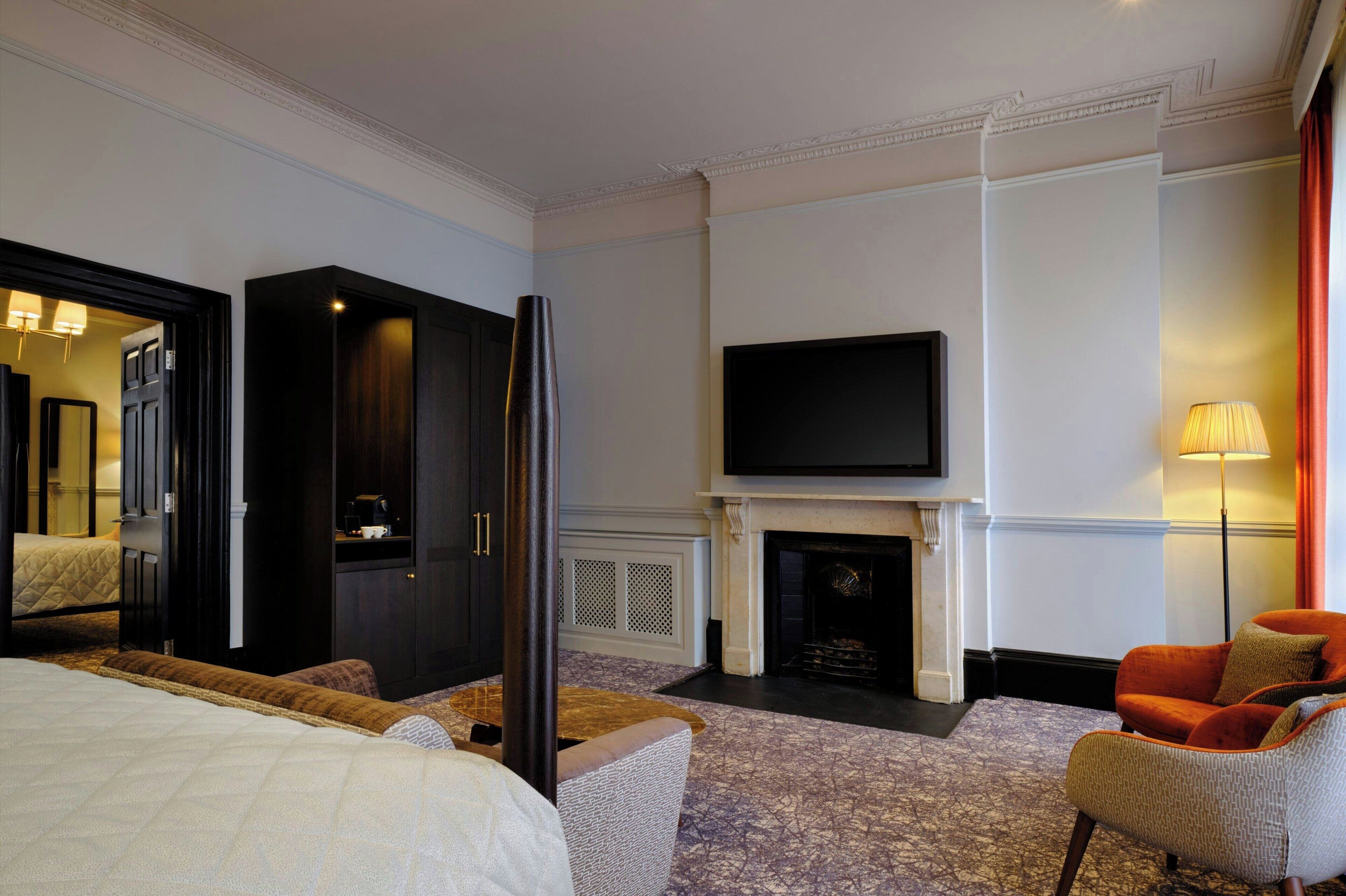 Une chambre d’hôtel comprenant un lit, un téléviseur fixé au-dessus d’une cheminée, un coin salon avec un fauteuil et un grand miroir.
