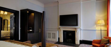 Une chambre d’hôtel comprenant un lit, un téléviseur fixé au-dessus d’une cheminée, un coin salon avec un fauteuil et un grand miroir.