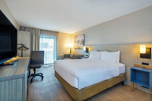 A hotel room with a bed, desk, chair, TV, lamp, and artwork on the wall.