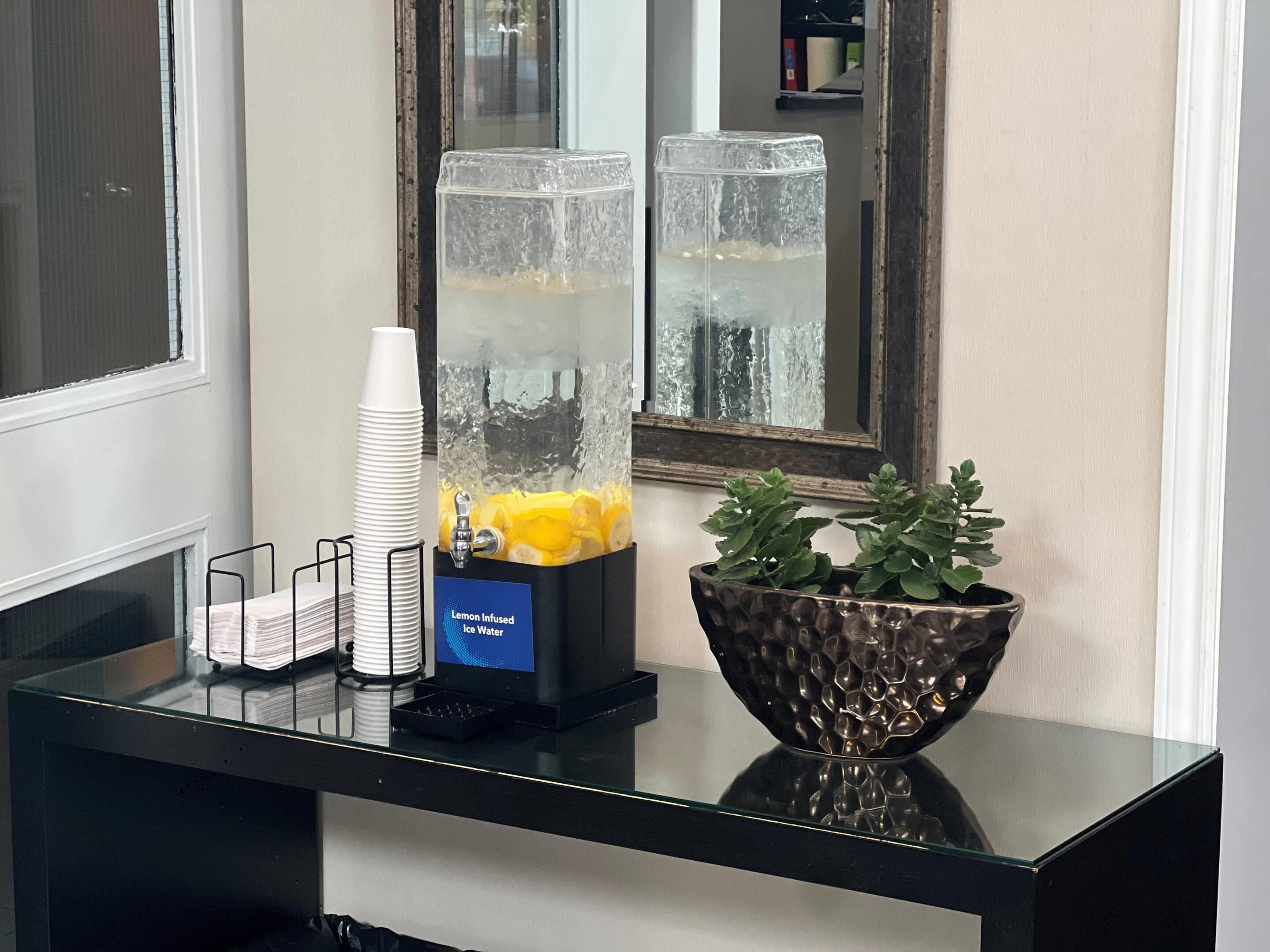 Een drankautomaat met water met citroen, een stapel papieren bekers en een decoratieve schaal met een plant.