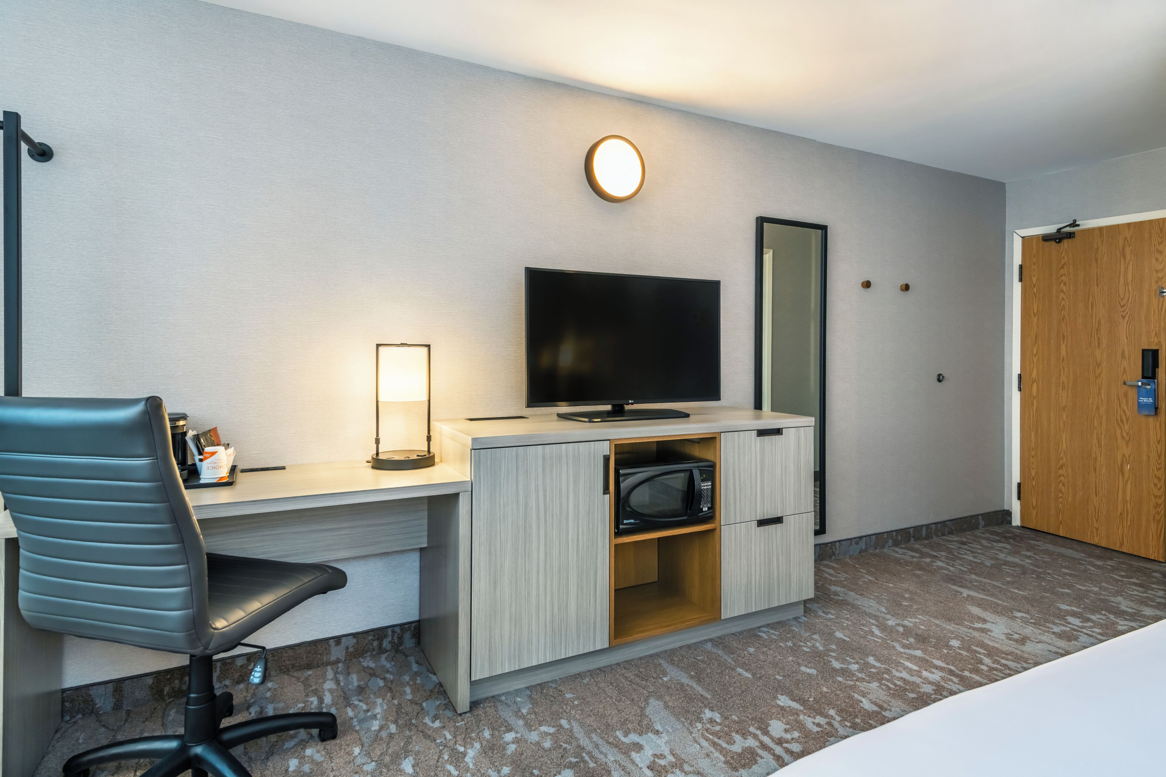 A hotel room with a desk, a chair, a television, a microwave, a bedside table, and a wooden door.