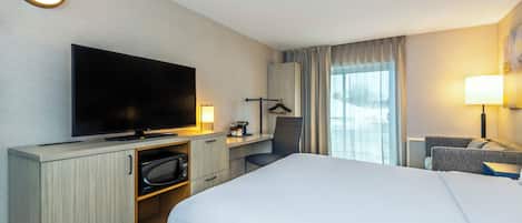 A hotel room with a large bed, a flat-screen TV, a desk with a chair, a coat rack, and a window with curtains.