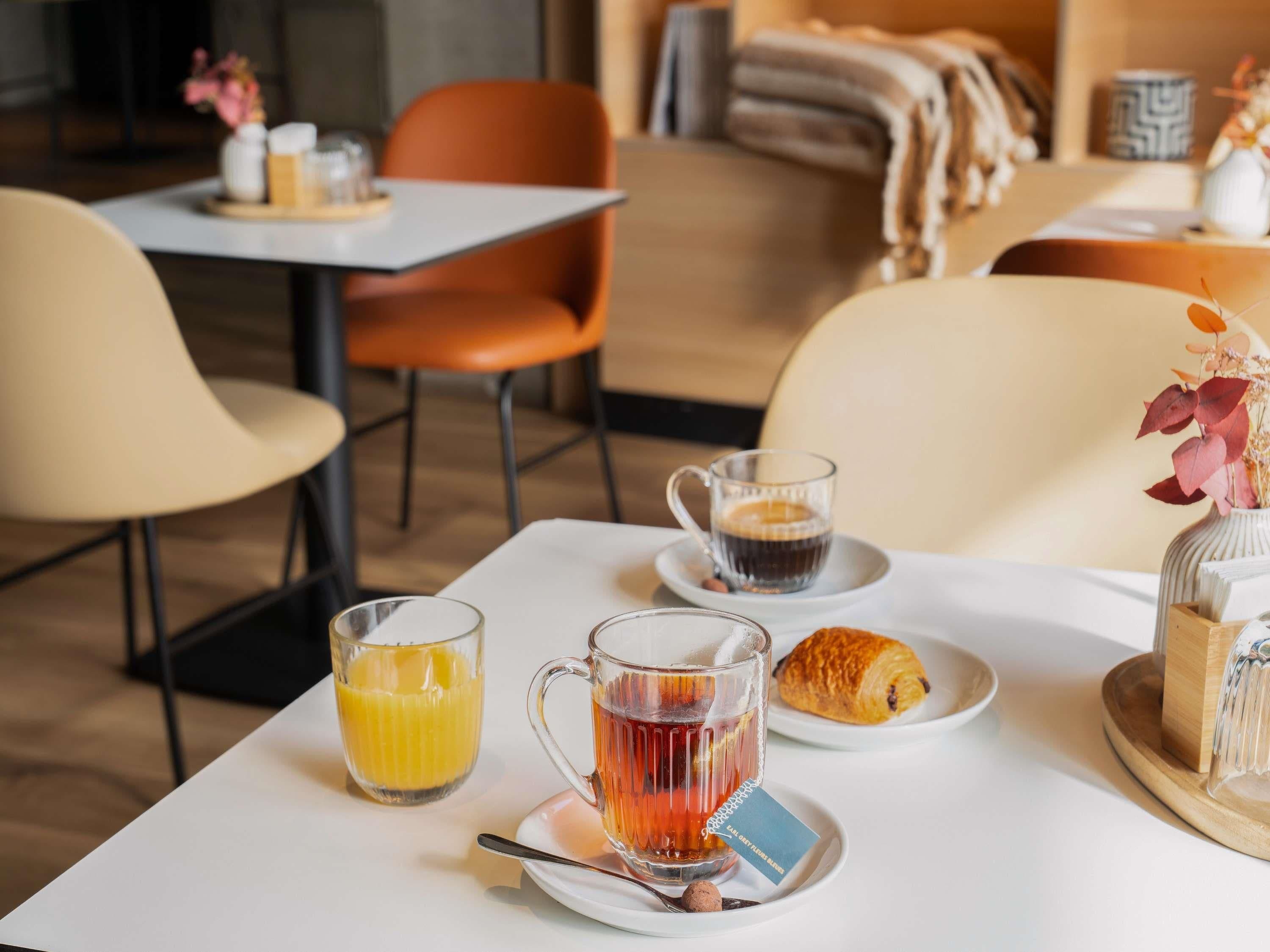A breakfast setting with a table, chairs, a coffee cup, a glass of orange juice, a teapot, a croissant, and a decorative vase.
