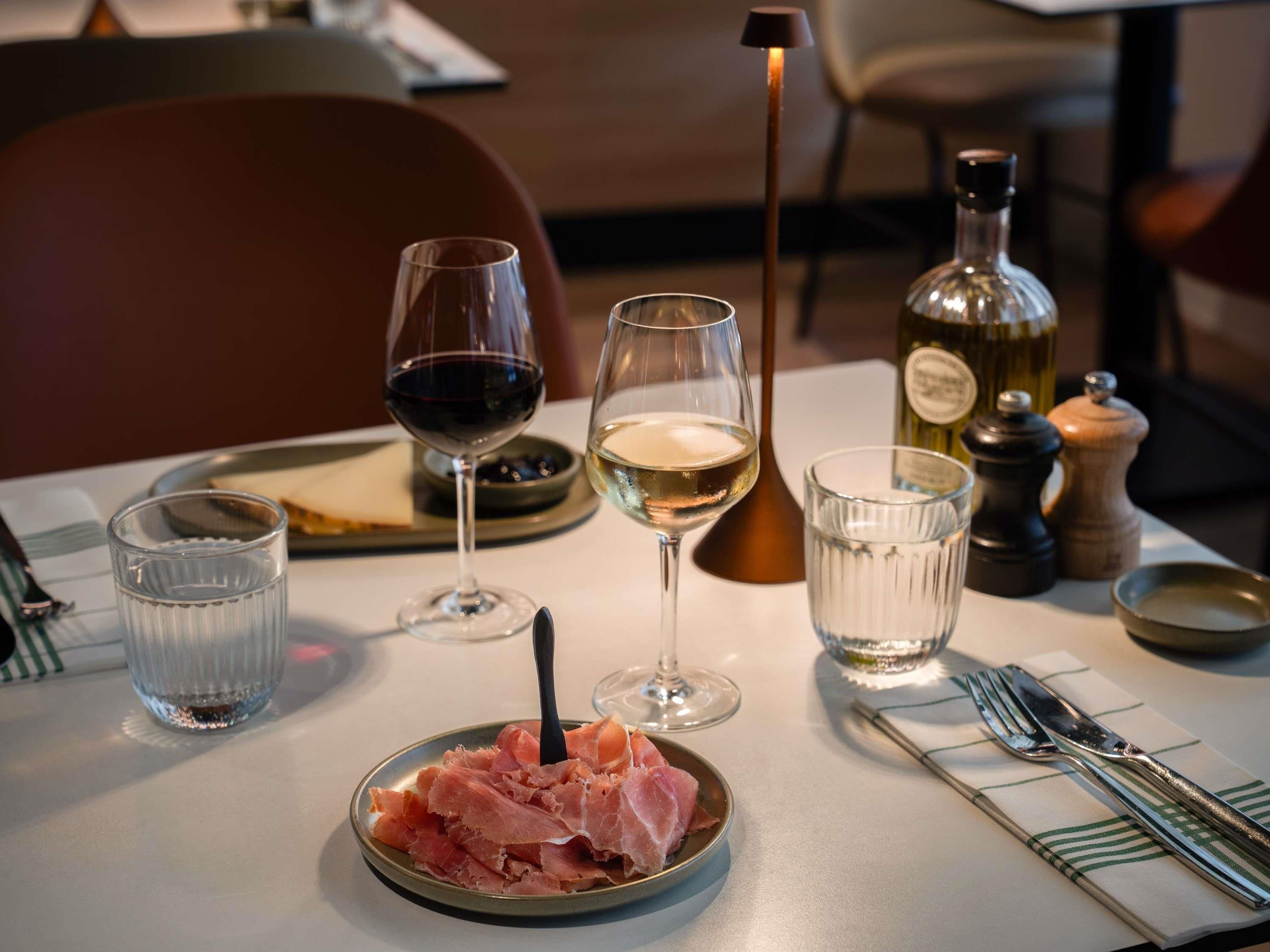 A dining table with a plate of cured meat, two glasses of wine, a bottle of olive oil, and a salt and pepper shaker.