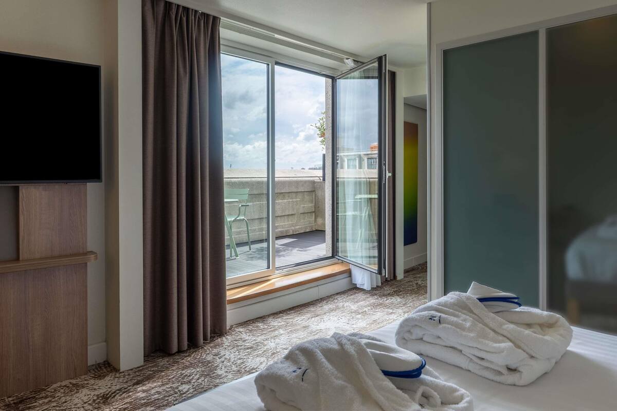 A hotel room with a large sliding glass door leading to an outdoor balcony, a flat-screen TV mounted on the wall, and neatly folded towels on the bed.