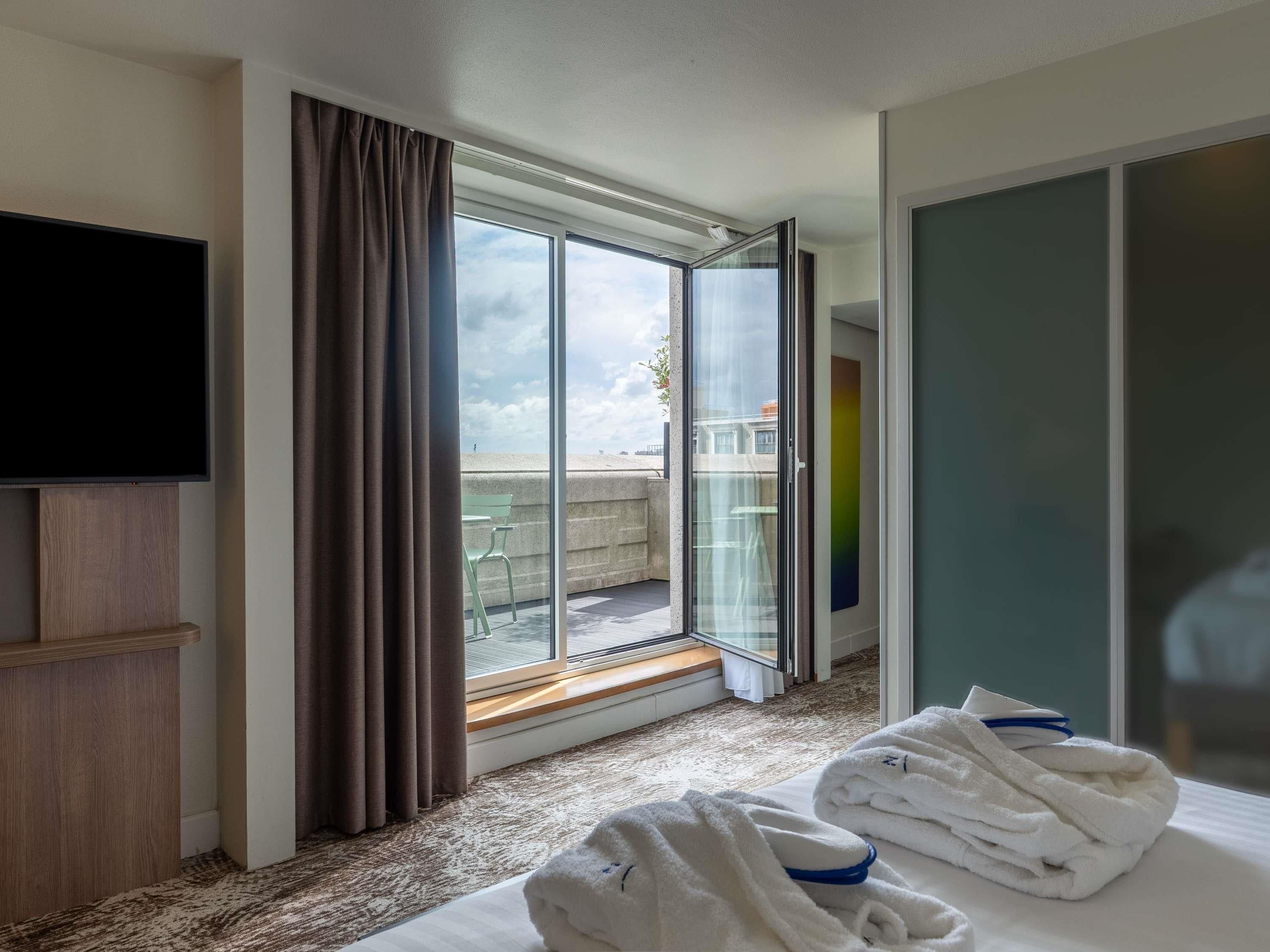 A hotel room with a large bed, a flat-screen TV, a sliding glass door leading to a balcony, and a set of white towels on the bed.
