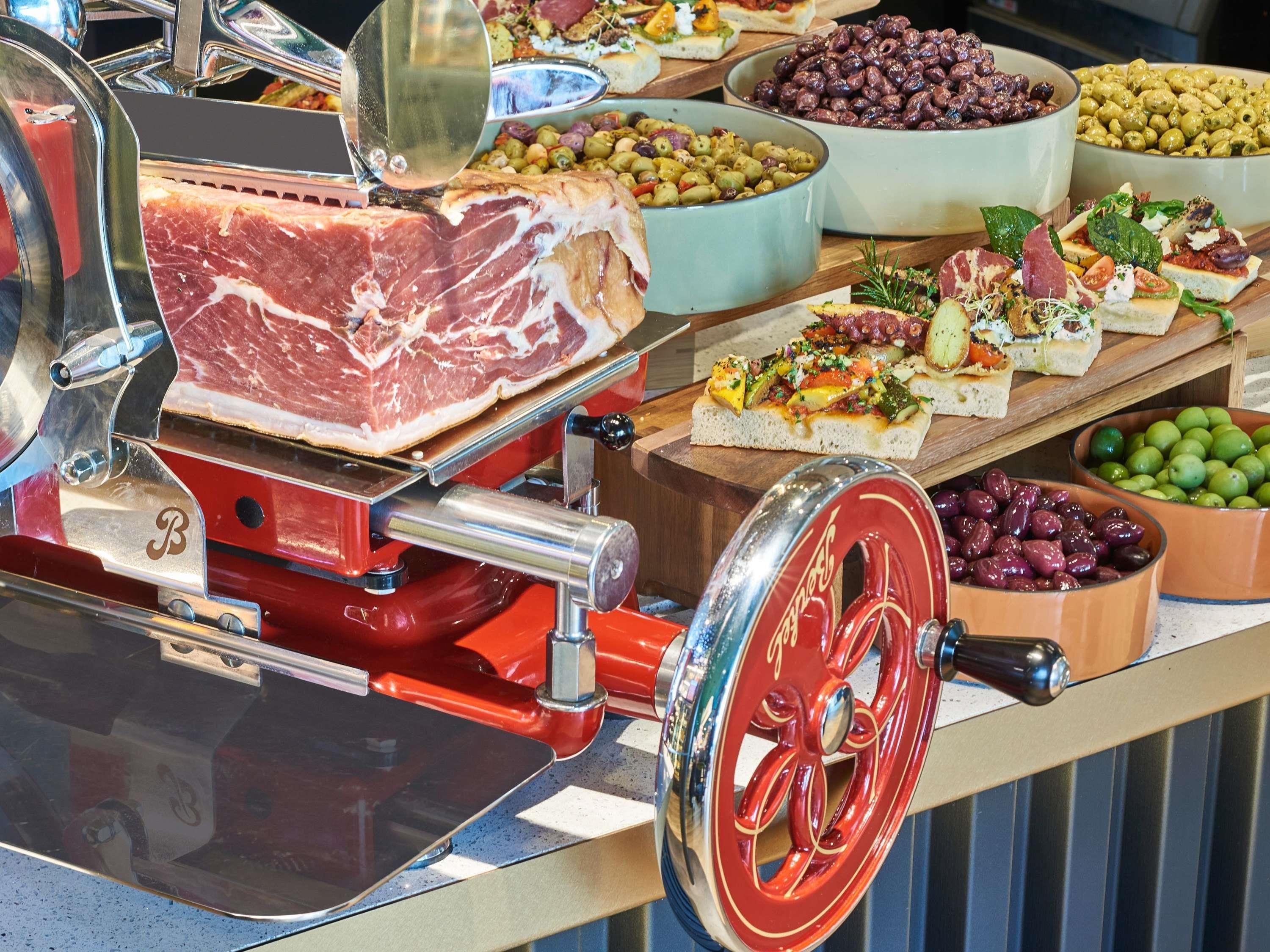 A meat slicer with a large piece of meat, bowls of olives, and various prepared dishes on a counter.