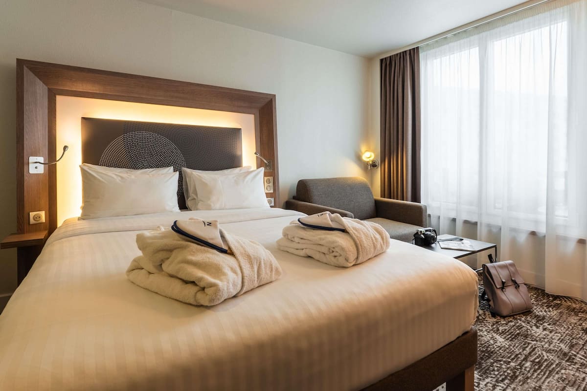 A hotel room with a large bed, a chair, a desk, and a window with curtains.