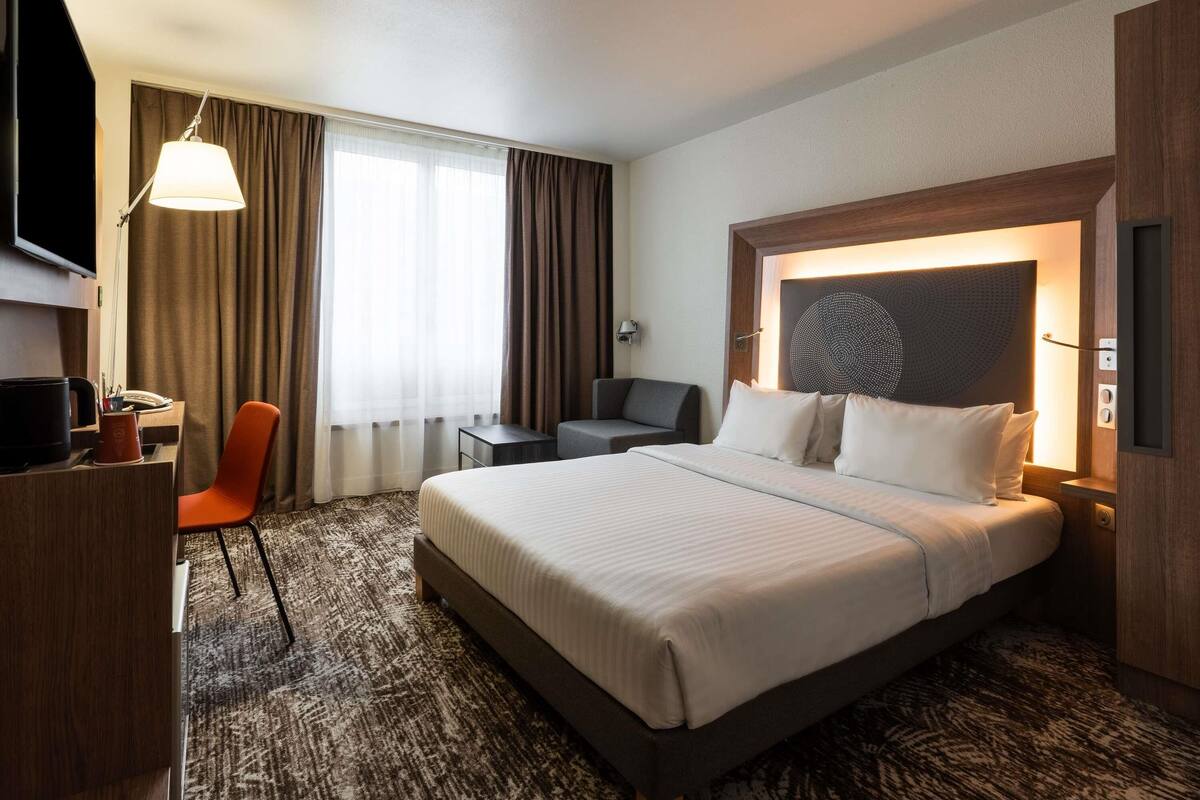 A hotel room with a large bed, a desk with a chair, a television, and a wall-mounted lamp.
