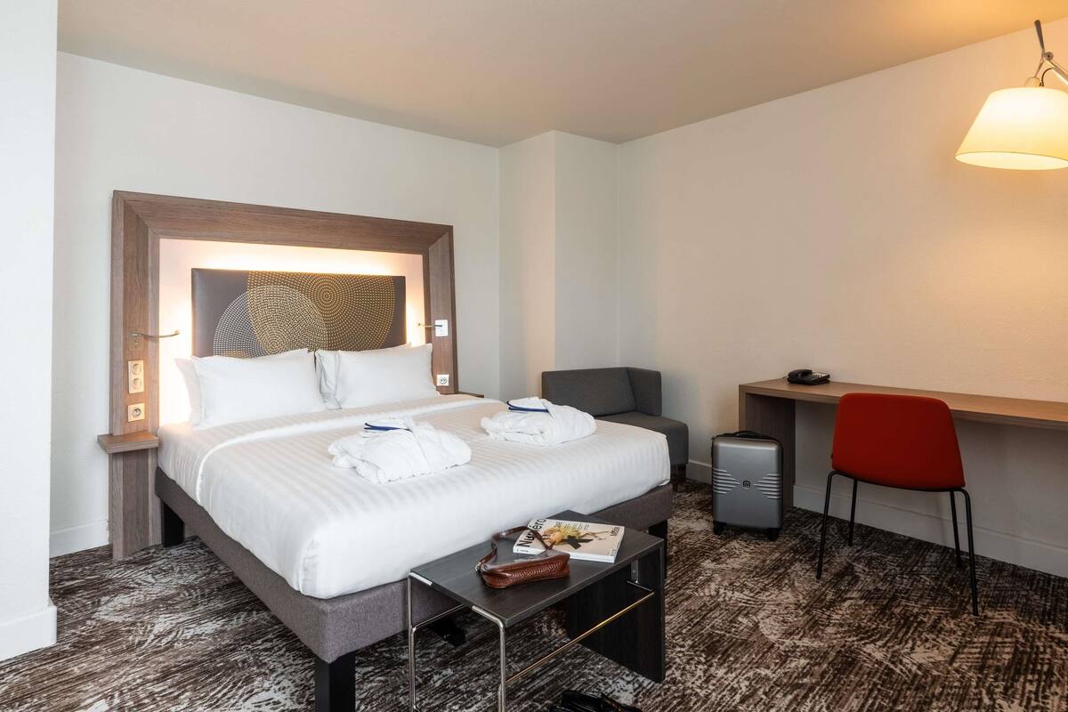 A hotel room with a bed, bedside table, desk, chair, and a lamp.