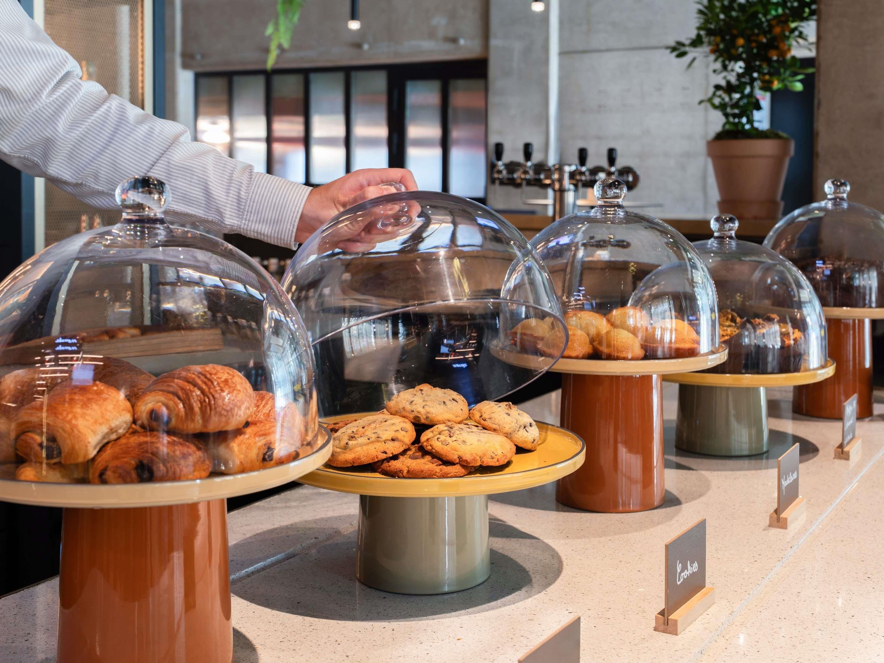 Eine Bäckertheke mit verschiedenen Backwaren unter Glassglocken, eine Person greift nach einem Gebäckstück, und ein Schild weist auf den Bereich hin.