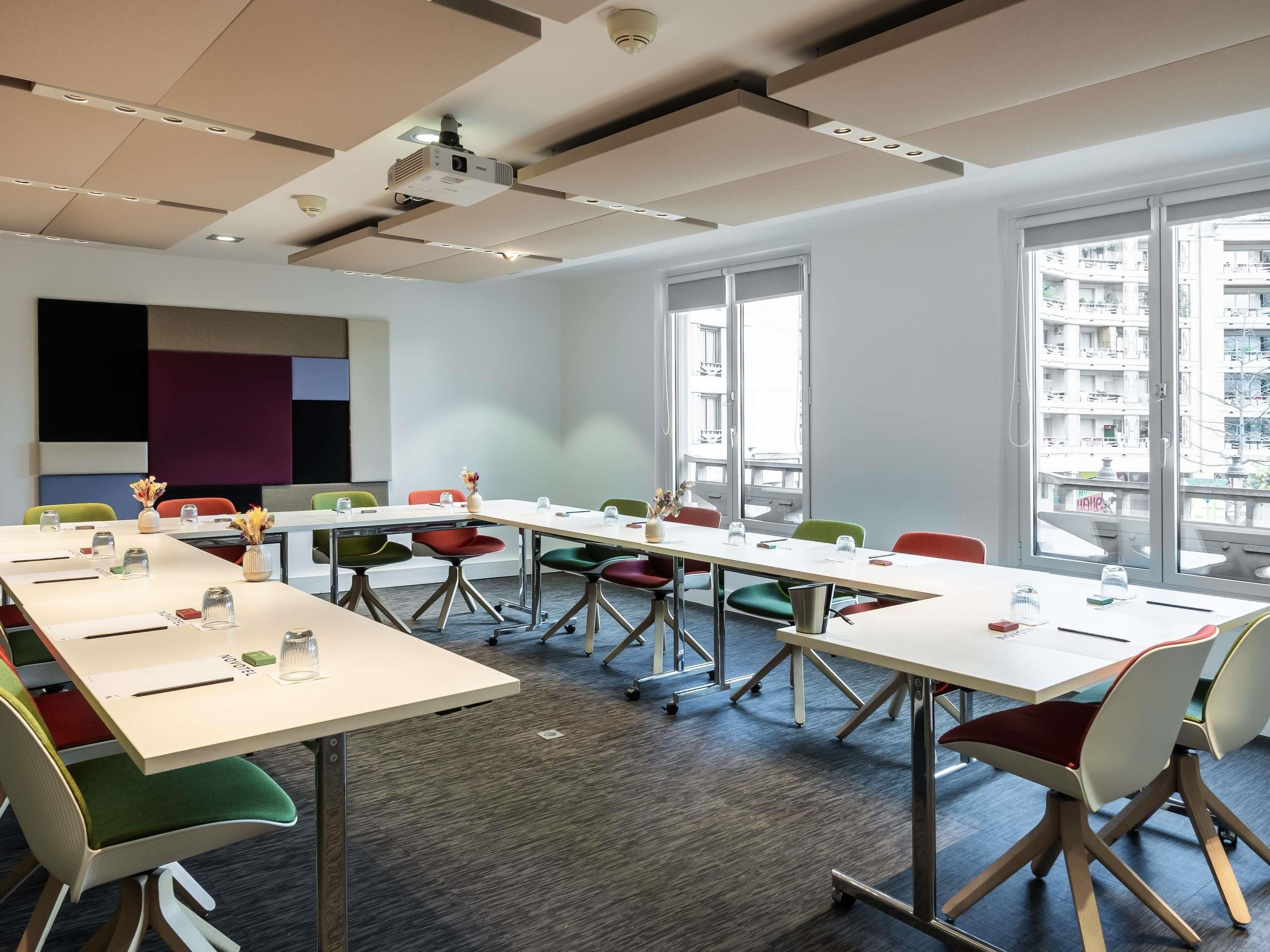 Sala de conferências com uma mesa comprida, cadeiras coloridas, um projetor e grandes janelas com vista para a cidade.