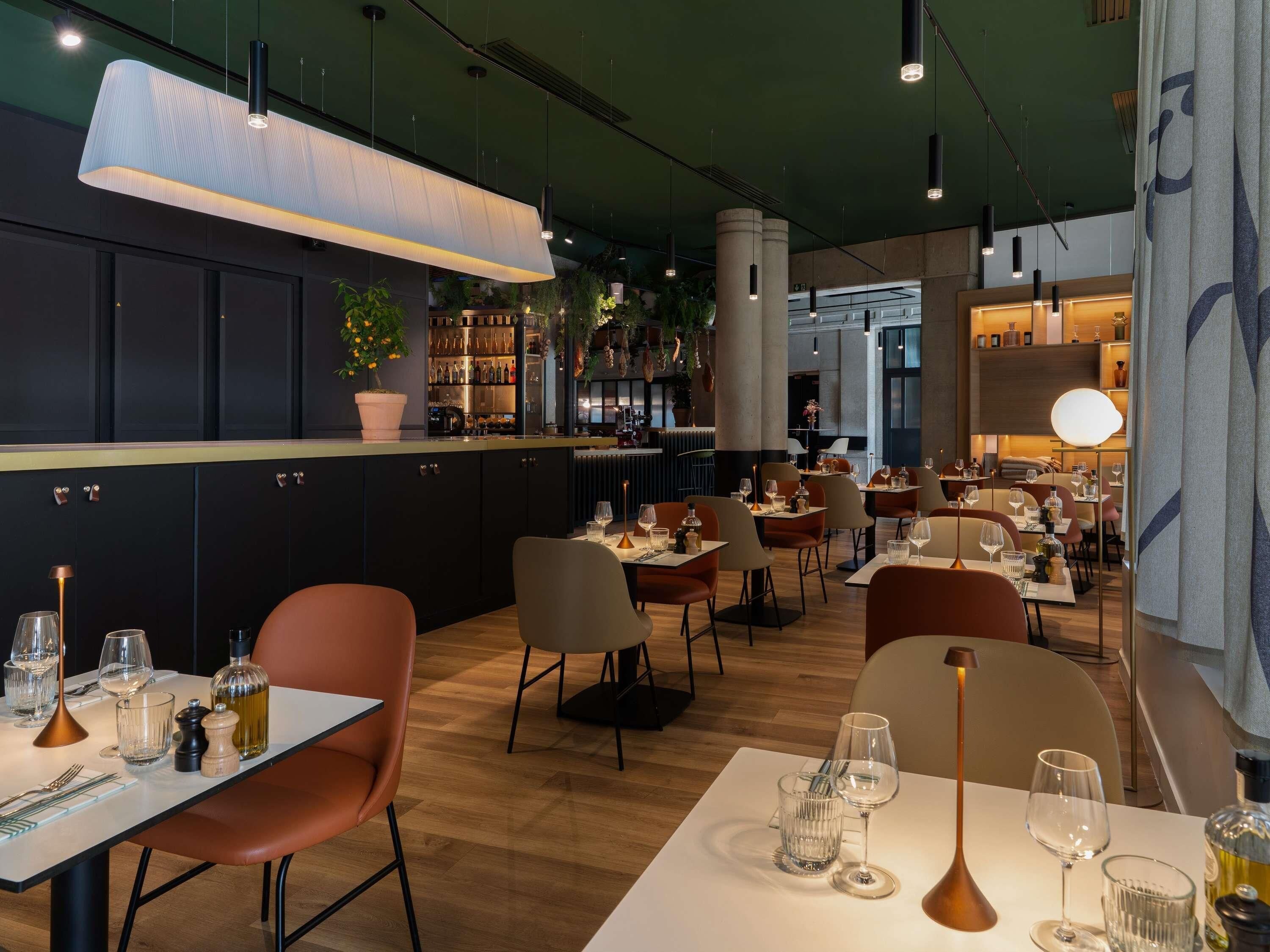 Interior de restaurante con mesas, sillas, zona de bar y luces colgantes.