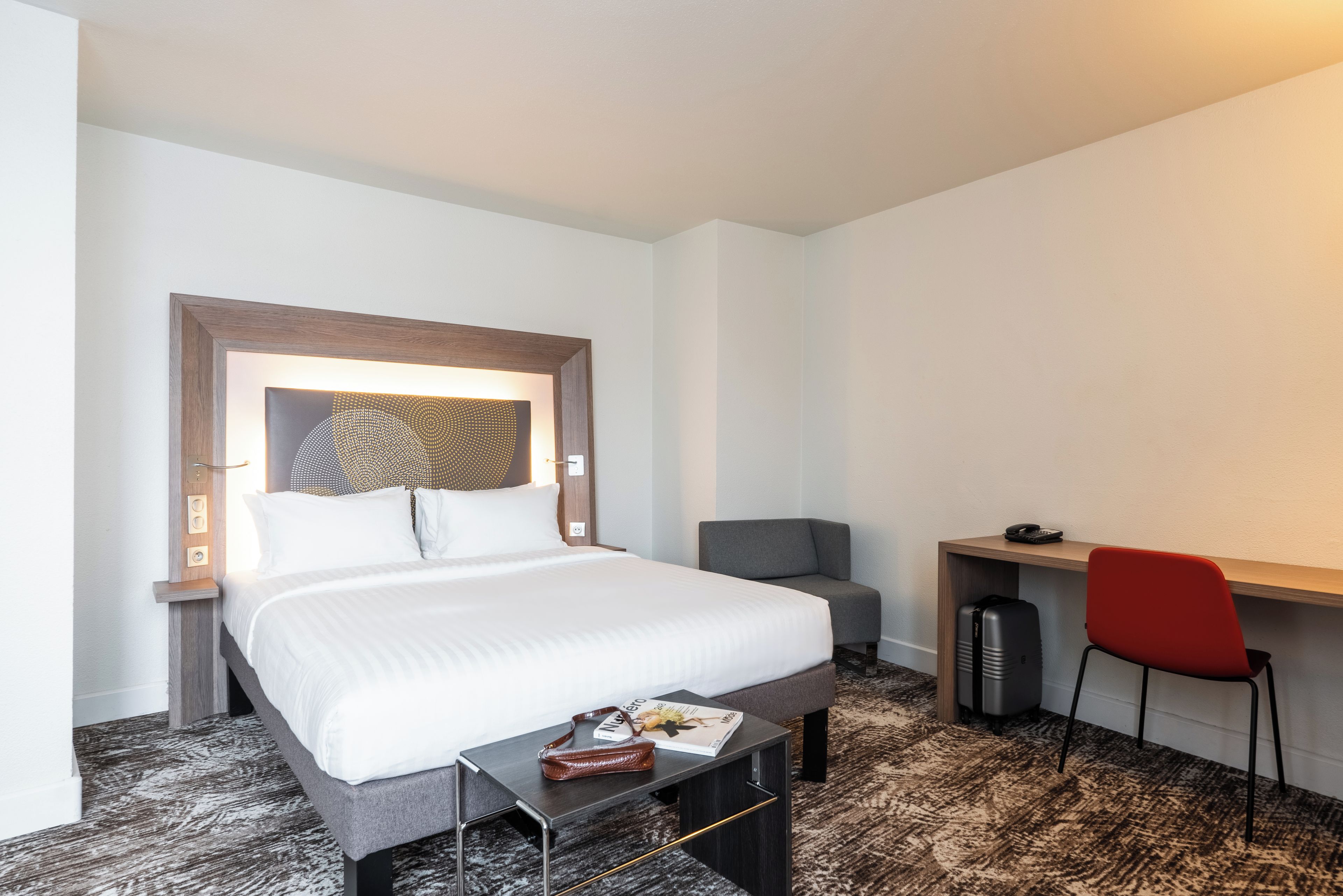 A hotel room with a bed, a desk, a chair, and a small table with a magazine.