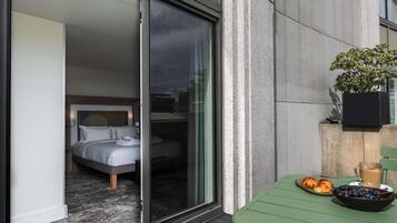 A modern hotel room with a sliding glass door leading to a balcony. The room features a bed with white linens and a headboard with a geometric pattern. A small table with a bowl of fruit and a cup of coffee is visible on the balcony.