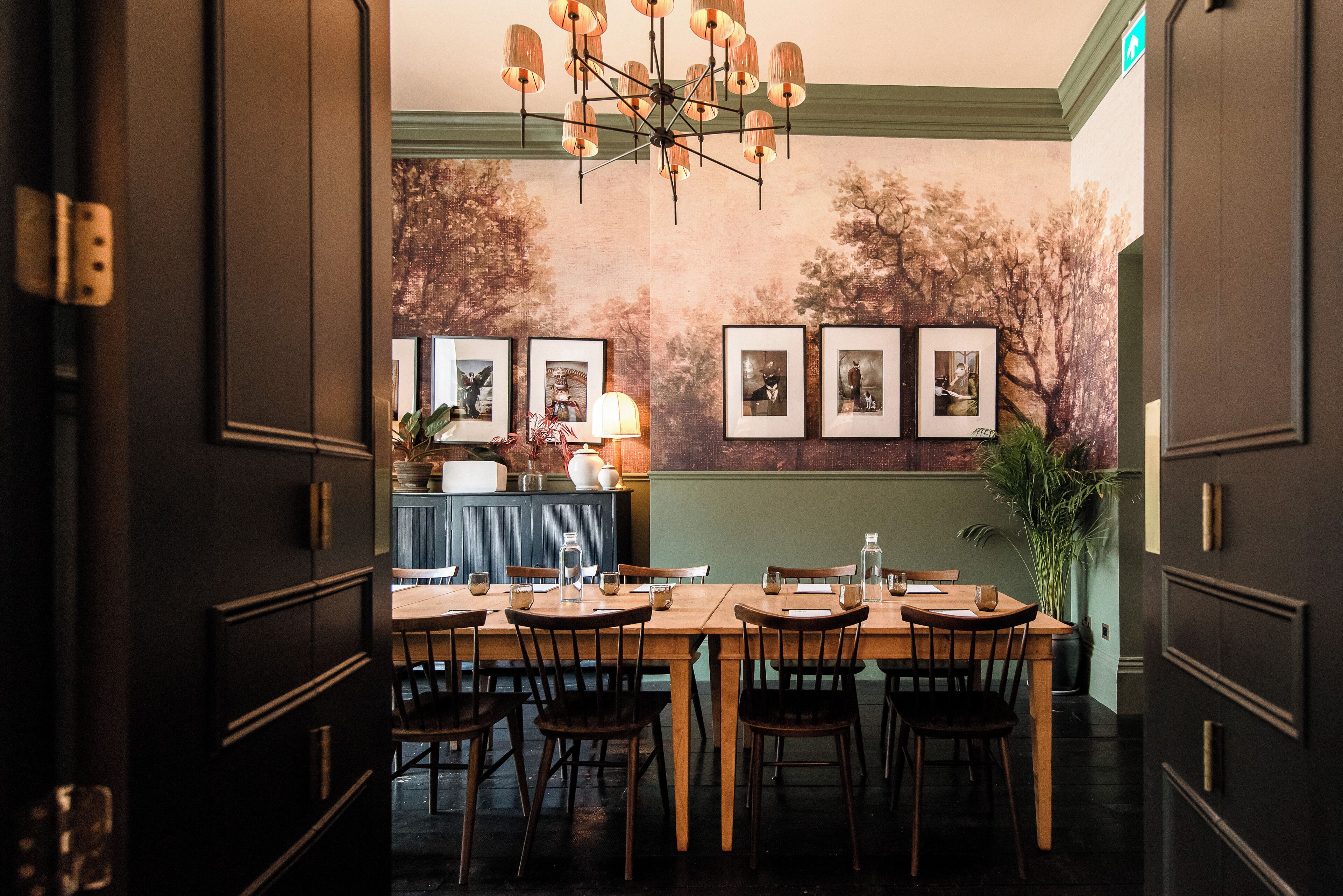 A dining area with a long wooden table, chairs, and a chandelier. There are framed pictures on the wall and a potted plant on the side.