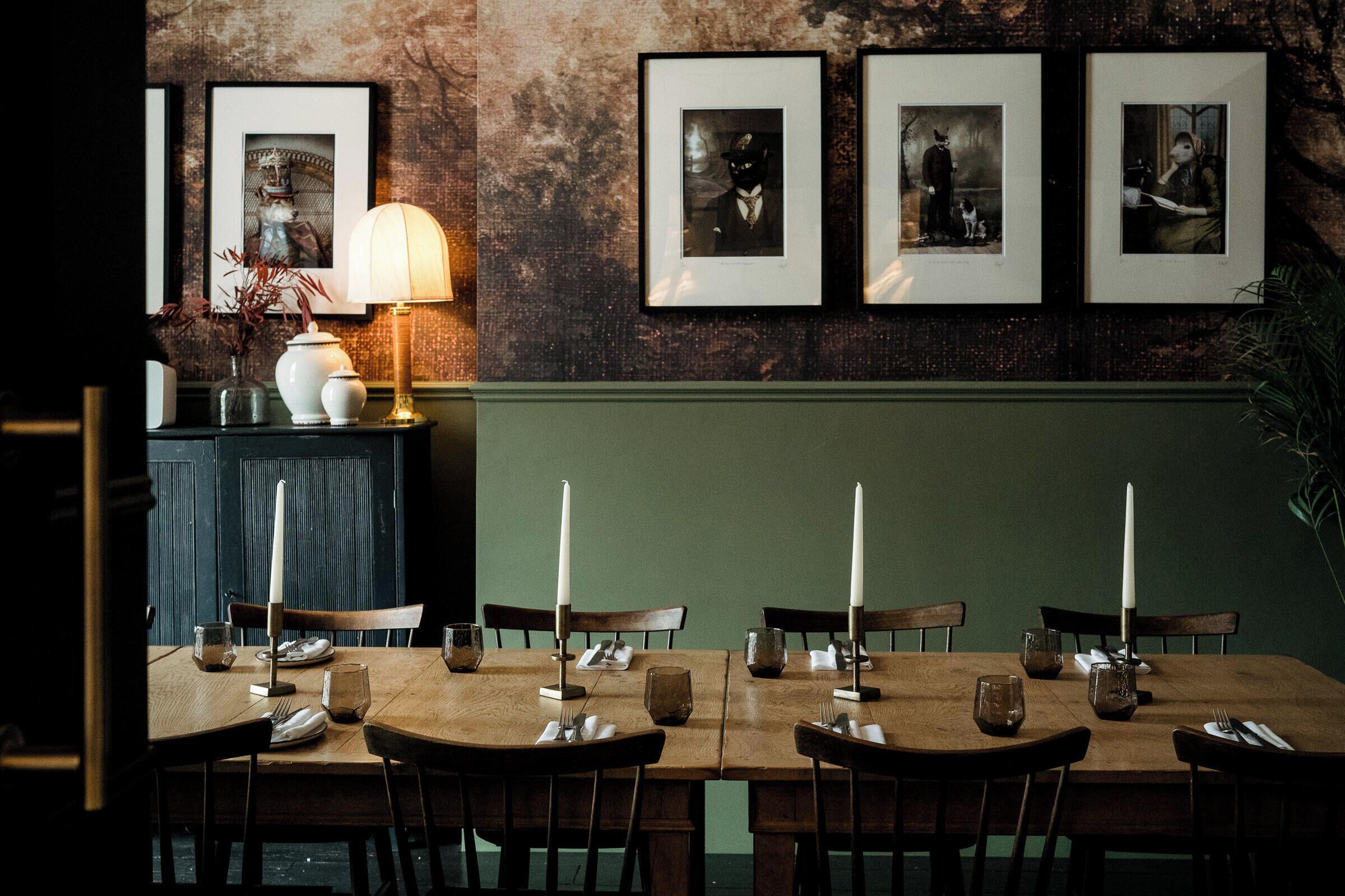 A dining area with a wooden table set for four, framed pictures on the wall, and a lamp on the sideboard.