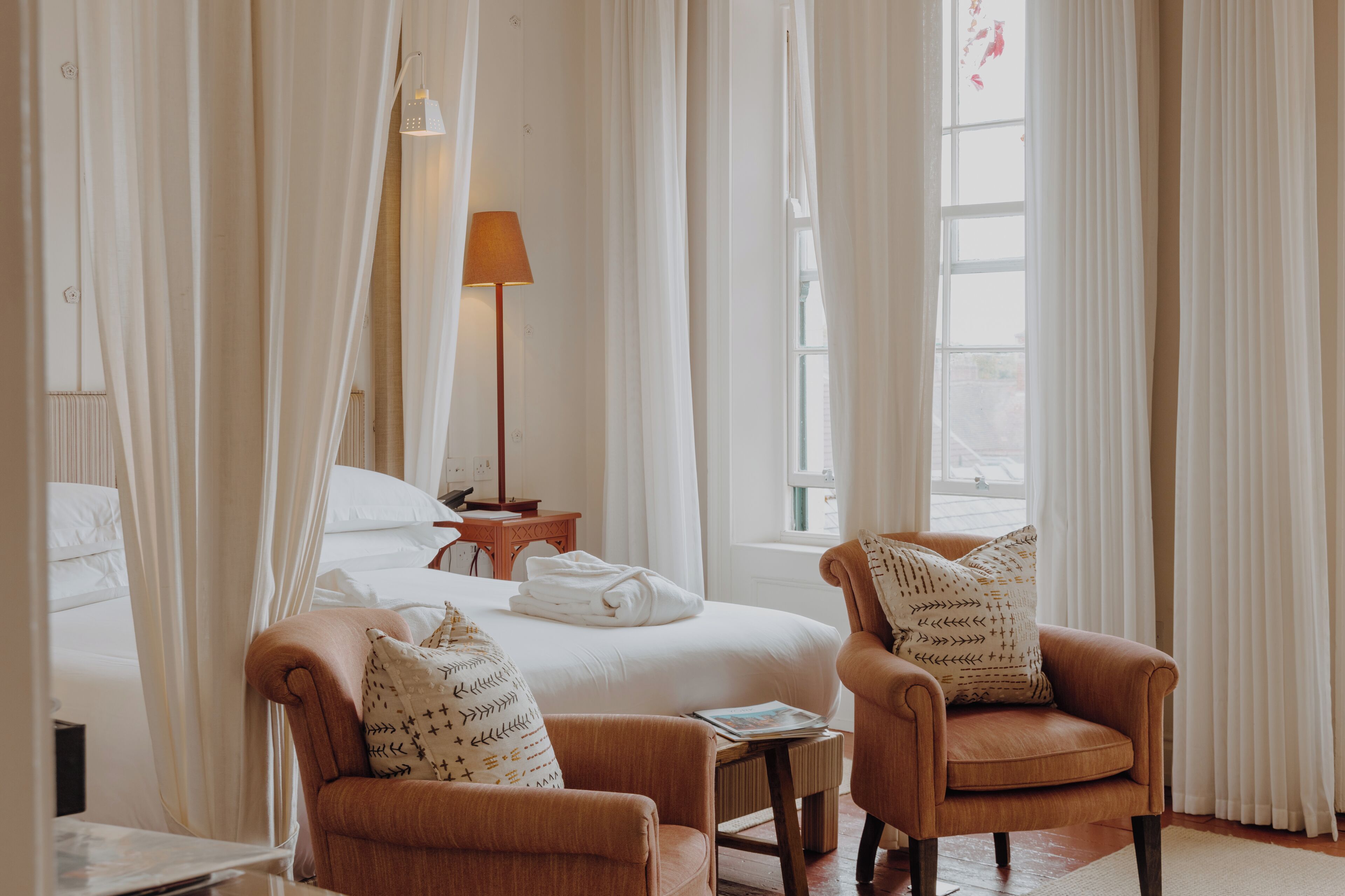 A hotel room with a bed, two armchairs, a bedside table, a lamp, and sheer curtains.