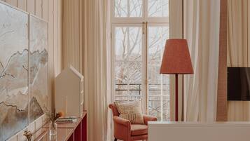 A room with a wooden floor, a glass table, a chair with a patterned cushion, a standing lamp, and a large window with curtains.