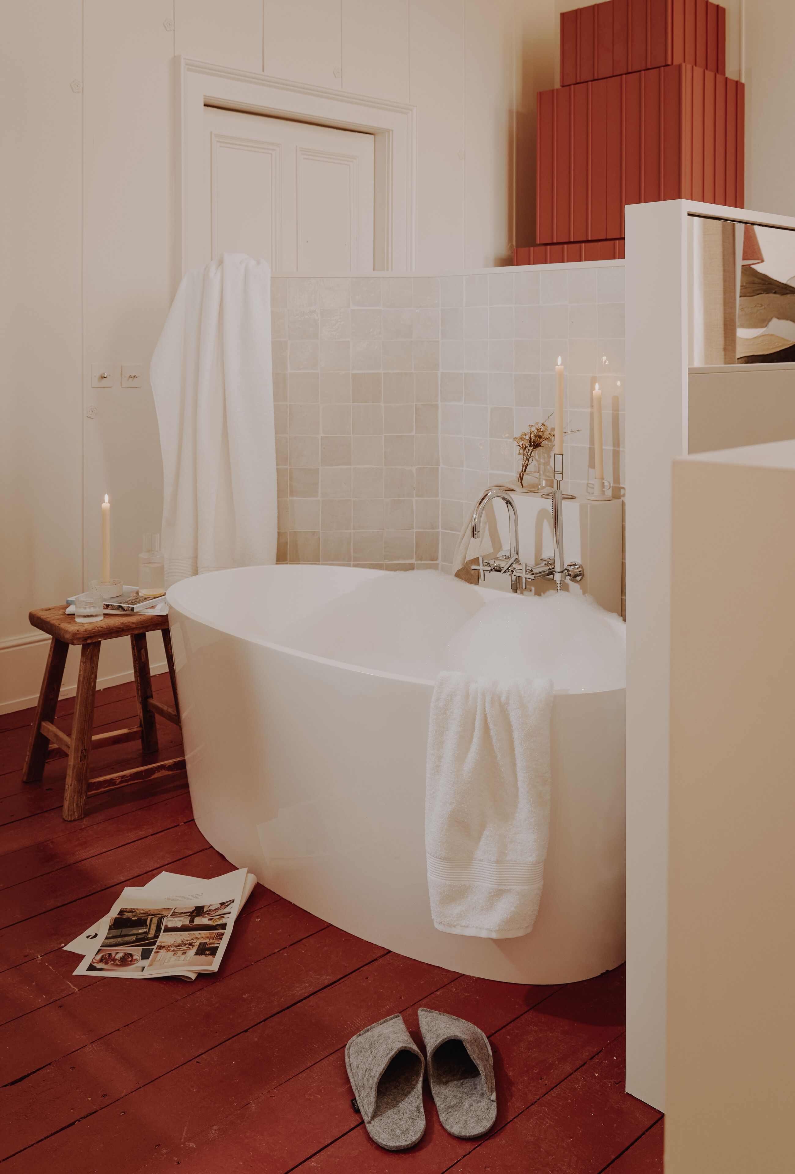 Ein modernes Badezimmer mit einer freistehenden Badewanne, einem hölzernen Hocker und Hausschuhen auf dem Boden.