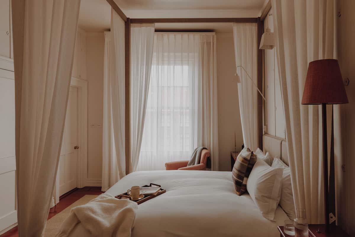 A hotel room with a large bed, a bedside table with a lamp, and sheer curtains.