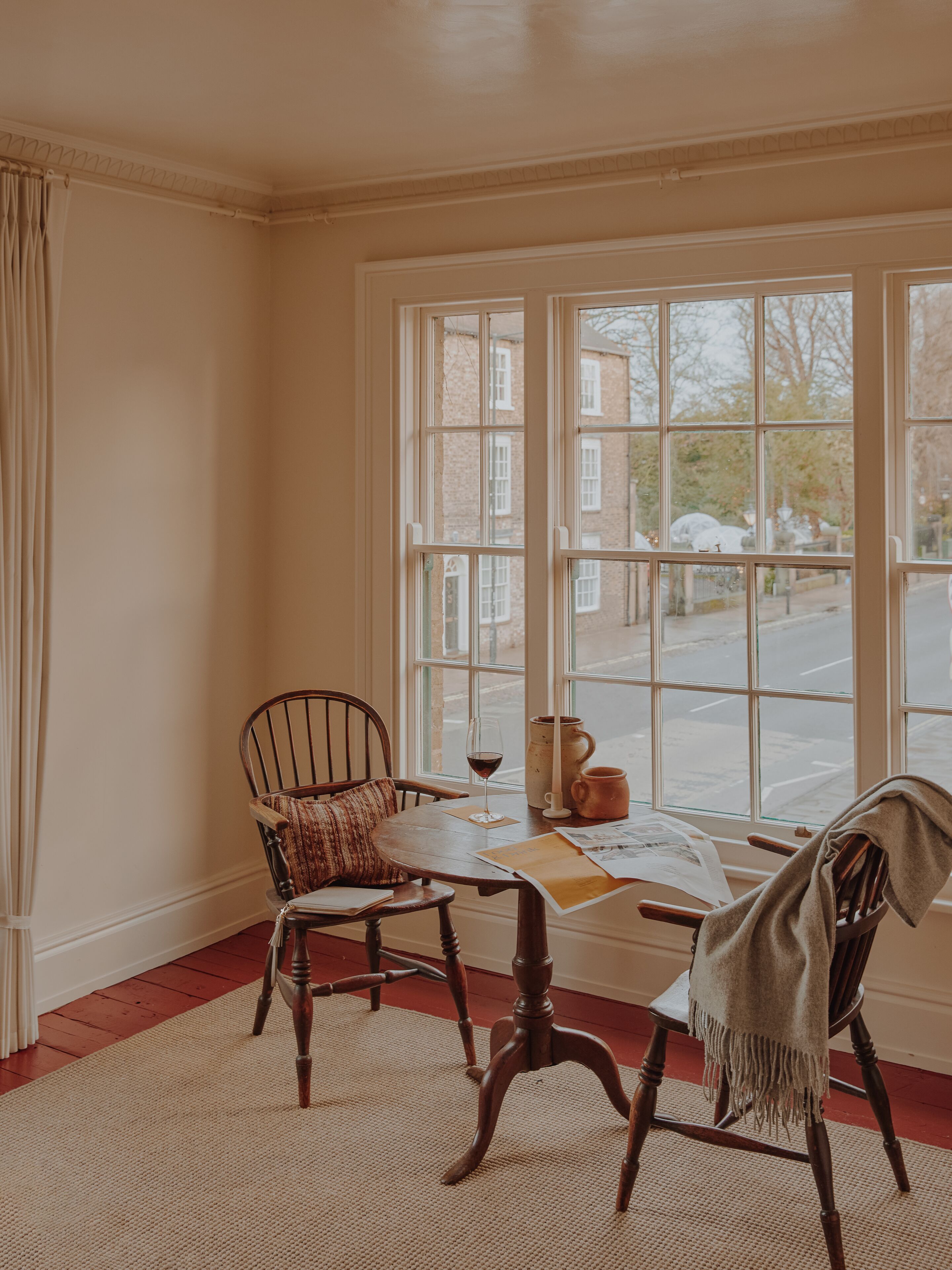 A room with a large window, a wooden chair, a table with a glass of wine, and a rug.