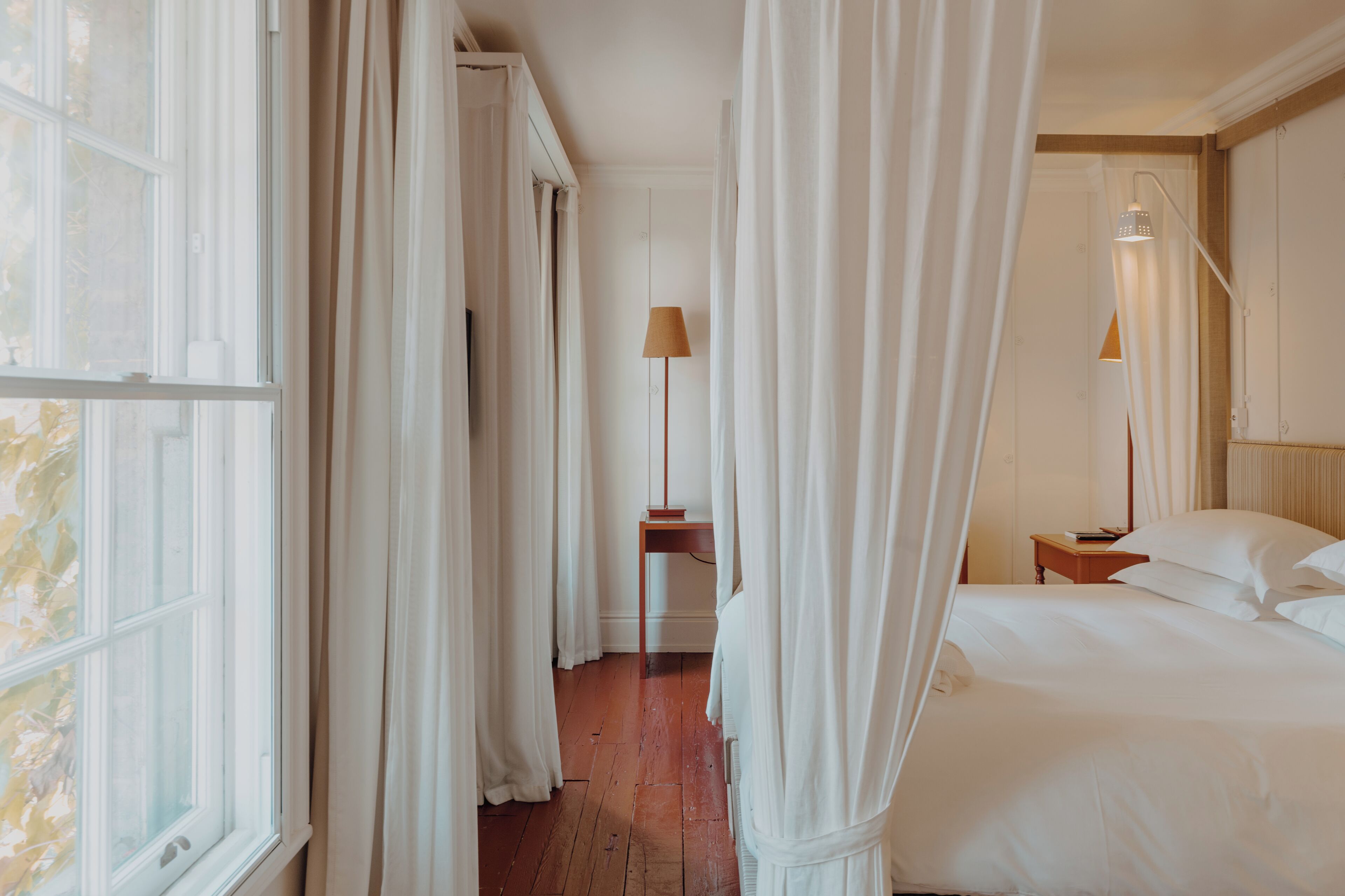 A hotel room with a wooden floor, a bed with white linens, a nightstand with a lamp, and sheer curtains.