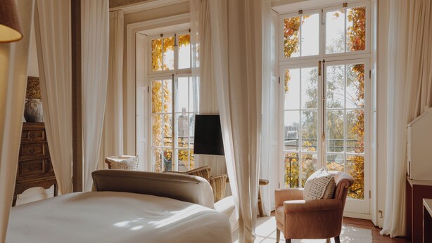 Une chambre avec un grand lit, une chaise, un téléviseur et une vue sur les feuilles d’automne à travers les fenêtres.