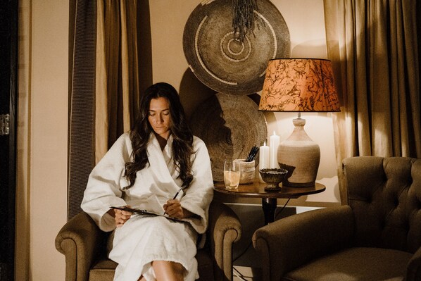 A person in a white bathrobe sitting on a chair, holding a pen and a notepad. There is a lamp with a patterned lampshade, a candle, and a small table with a glass and a bowl.